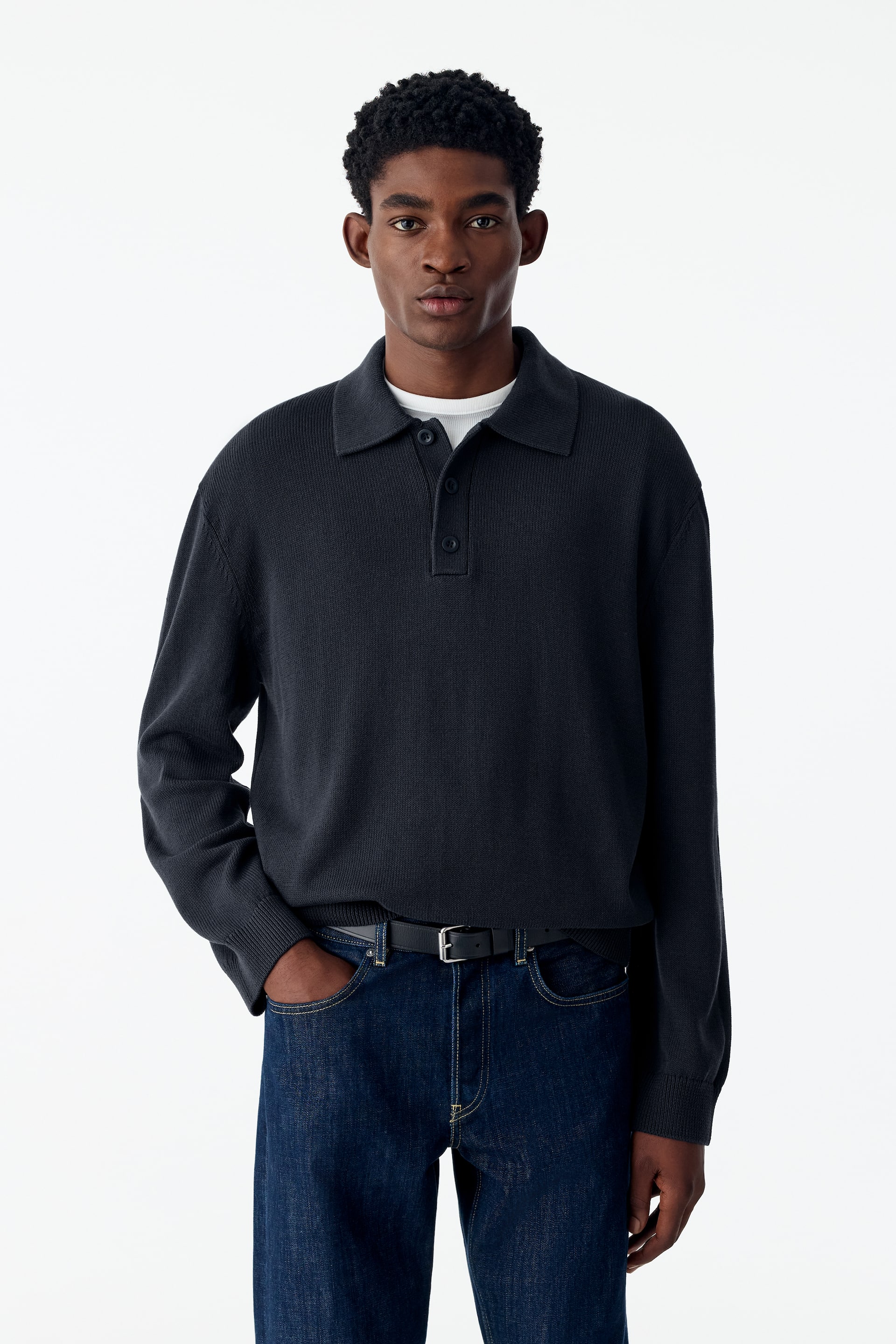 Ver imagem maior: O modelo veste uma polo de manga comprida em malha fina, cinzento-escura, sobre uma t-shirt branca e por dentro de uns jeans azuis escuros. A polo de corte descontraído apresenta gola clássica, carcela parcial com fecho de três botões e punhos canelados.
