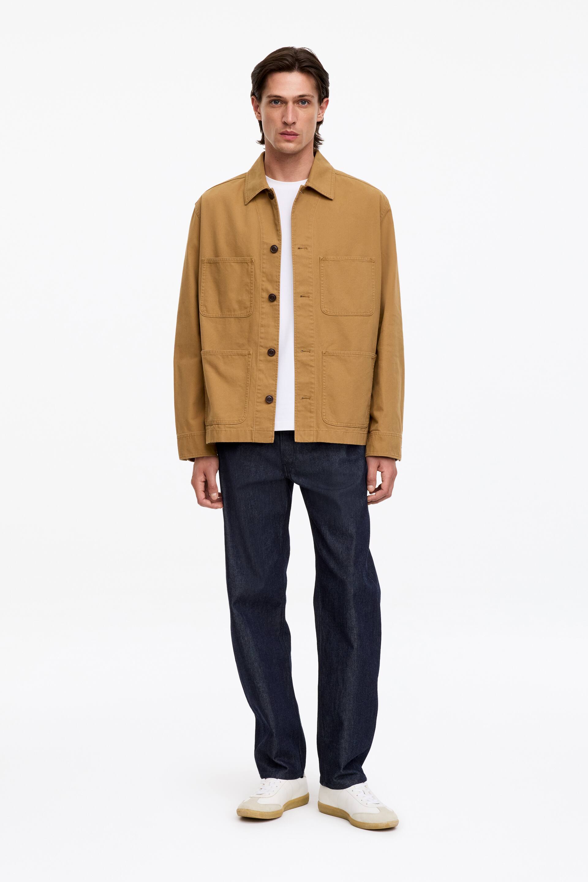 Se større billede: En brunlig utility-overshirt med klassisk krave og fire påsyede lommer foran bæres åben af modellen. Overshirten er stylet i lag over en hvid T-shirt og kombineret med mørkeblå jeans.