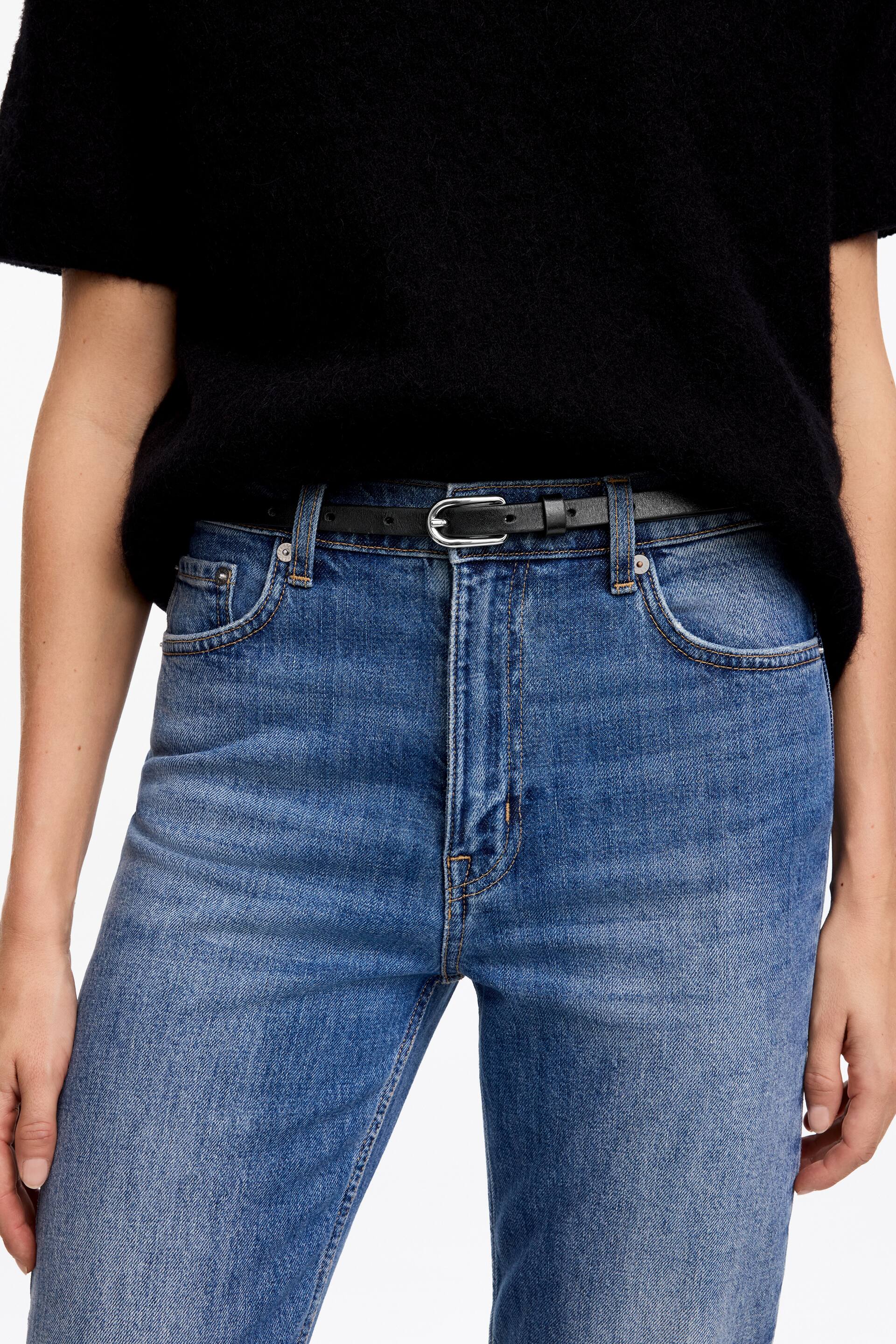 View larger image: A slim black leather belt with a silver-toned oval buckle is worn by the model through the loops of high-waisted blue jeans. The belt is styled with a tucked-in, short-sleeved black knitted top.