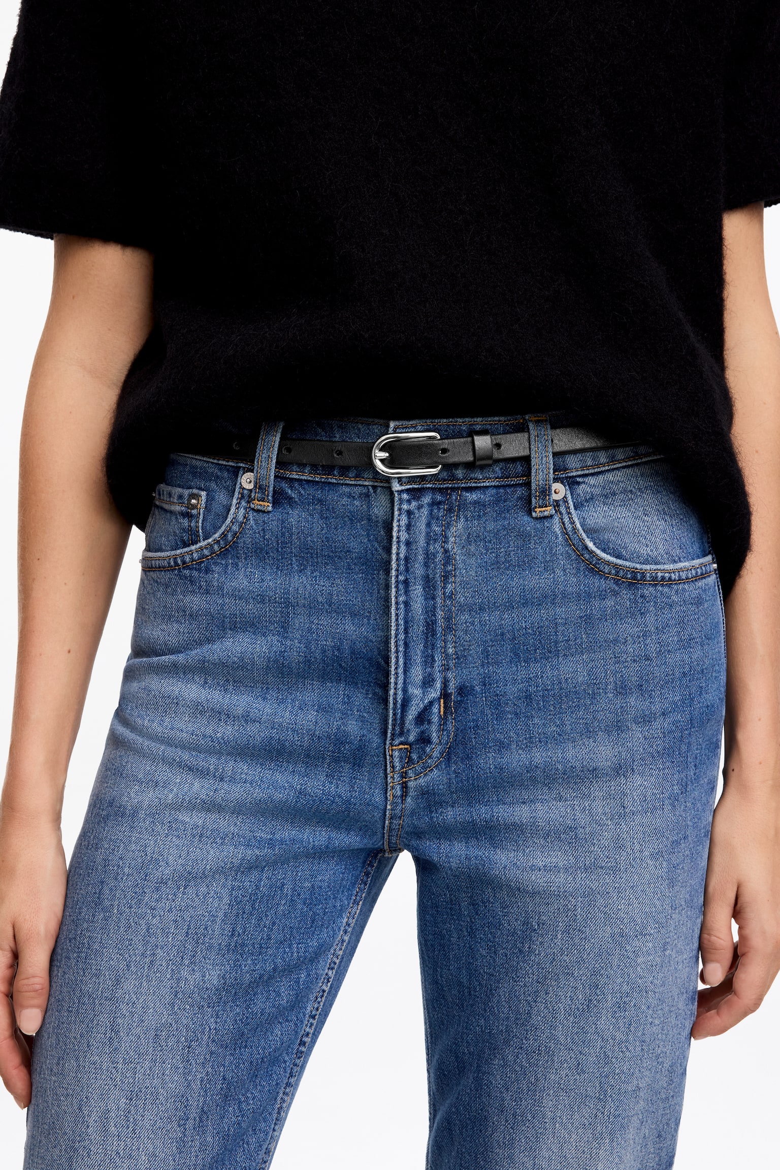 Ein schmaler schwarzer Ledergürtel mit einer ovalen Schnalle in Silberoptik wird vom Model durch die Gürtelschlaufen einer hochgeschnittenen blauen Jeans getragen. Der Gürtel ist mit einem in die Hose gesteckten schwarzen Strickoberteil mit kurzen Ãrmeln kombiniert. - 1