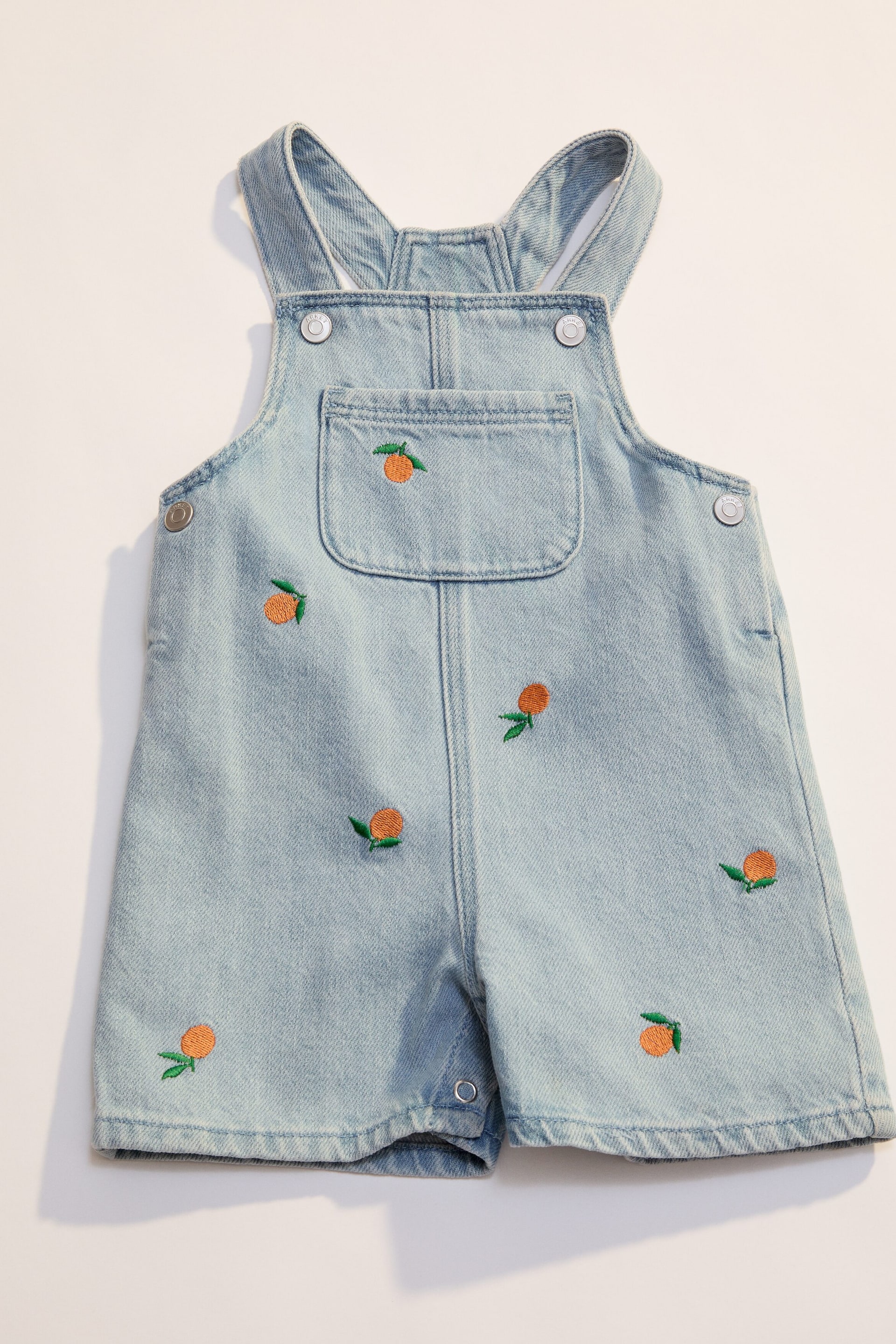 Grösseres Bild ansehen: Die Vorderseite der hellblauen Jeans-Latzshorts zeigt ein durchgehendes, gesticktes Orangenmuster. Die Shorts verfügen über Schulterträger, eine einzelne aufgesetzte Tasche auf dem Latz und Metallschnappverschlüsse an den Trägern und der Seite.