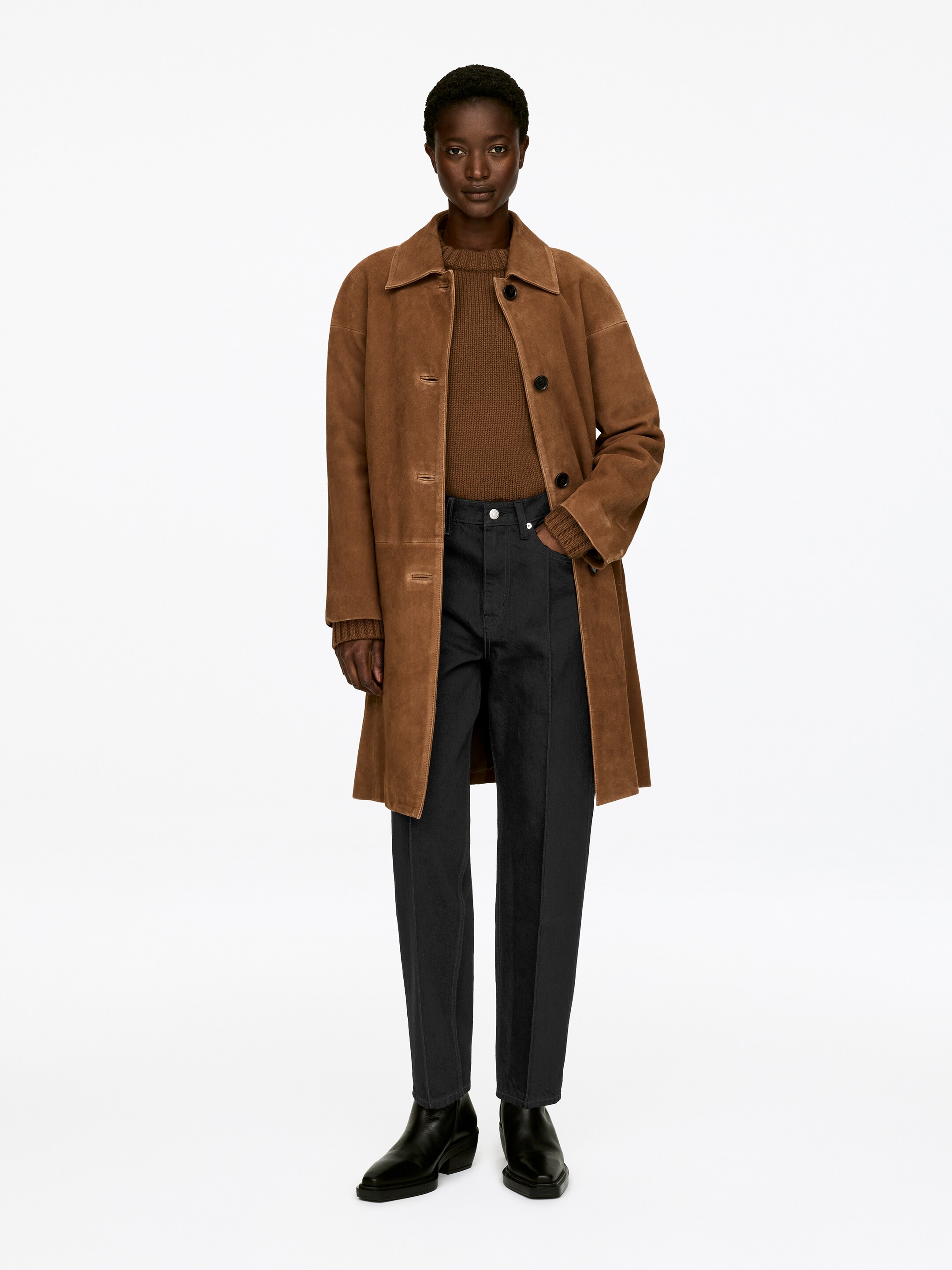 Agrandir l'image: Un manteau en daim marron est présenté en vue de face complète, avec un col pointu, des manches longues, une fermeture par boutons à l'avant, et atteignant le milieu de la cuisse. Il est superposé à un pull tricoté marron et un pantalon noir à jambes droites, qui présente une couture avant visible, et est stylé avec des bottines à bout pointu noires.
