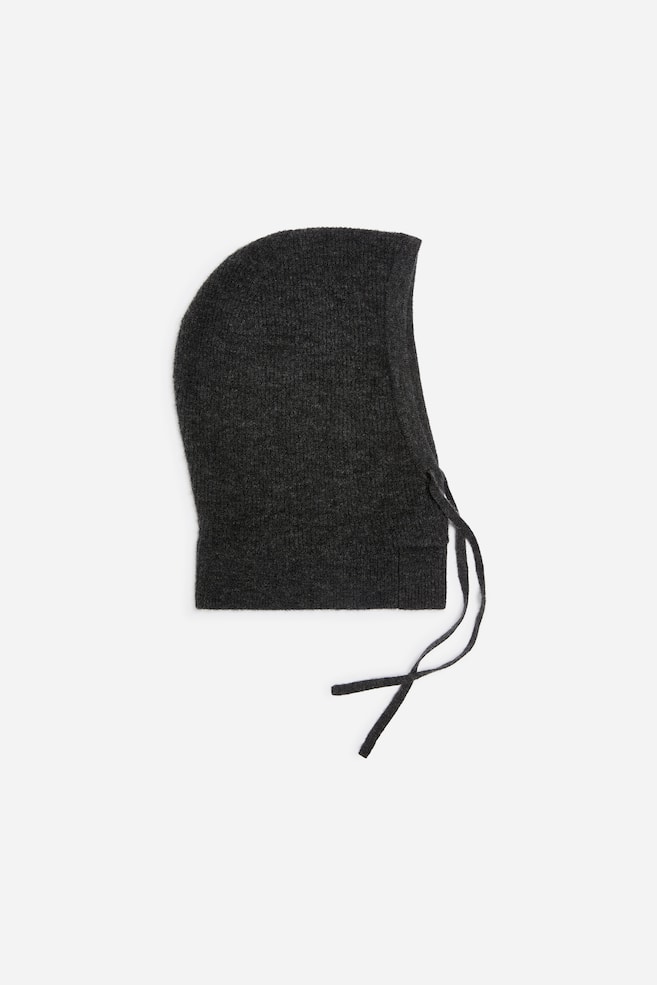 Eine dunkelgraue Strickkapuze ist im Profil nach rechts ausgerichtet und zeichnet sich durch ein weiches, strukturiertes Material, einen gerippten Nackenbund sowie dünne, farblich passende Strickbänder aus, die am vorderen Ausschnitt herabhängen. - 1