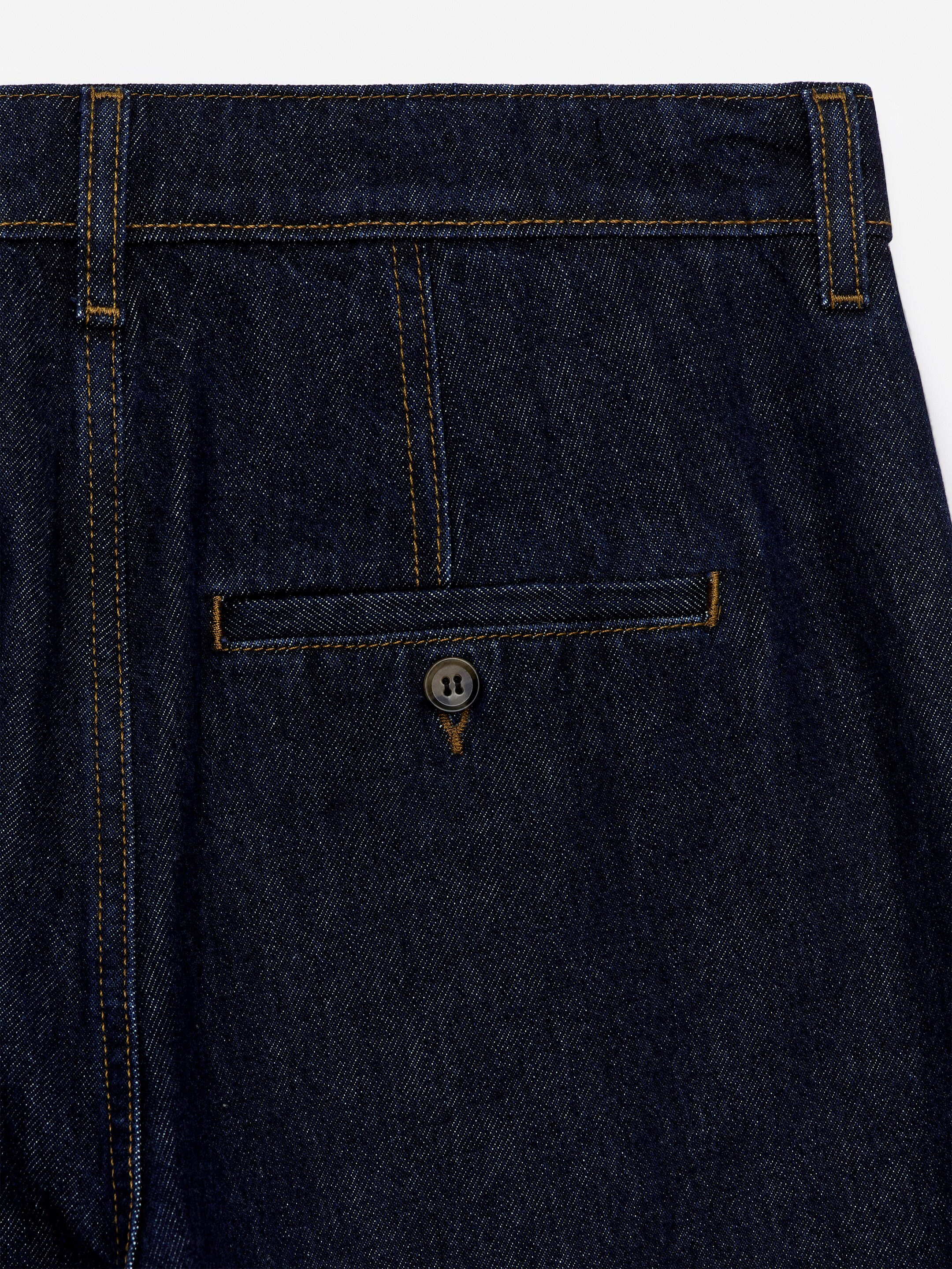 Grotere afbeelding bekijken: Donkerblauwe denim broeken tonen een gedetailleerd beeld van het bovenste ruggedeelte, met gouden contraststiksels langs de tailleband, naden en een klepzak. De zak is vastgemaakt met een donkere knoop met vier gaten.