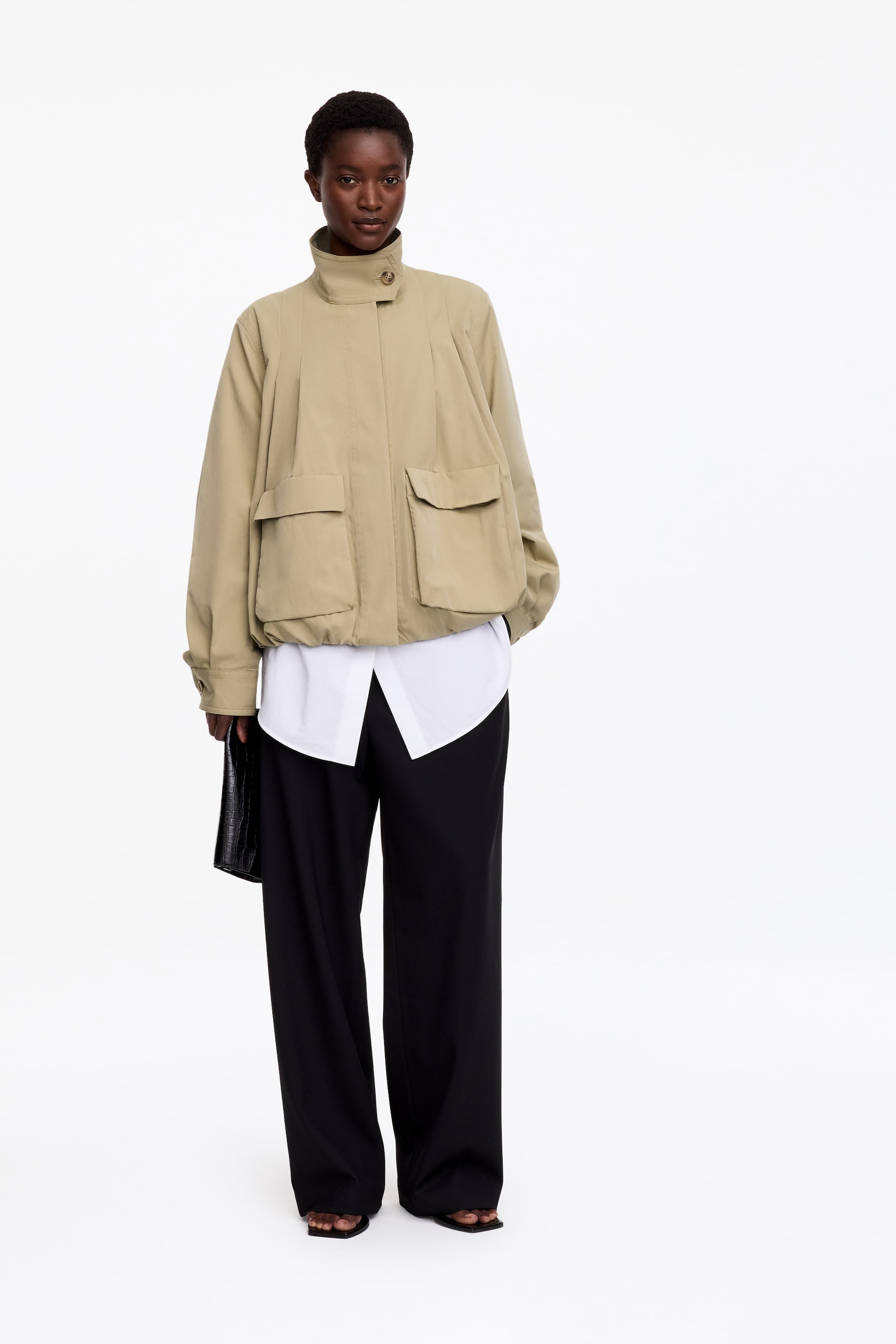 View larger image: A beige short blouson jacket is worn by the model, featuring a high stand collar, front pleat detailing, large flap pockets, and a softly rounded bubble hem. The jacket is layered over a white collared shirt and wide-leg black trousers, while the model is holding a black clutch bag.