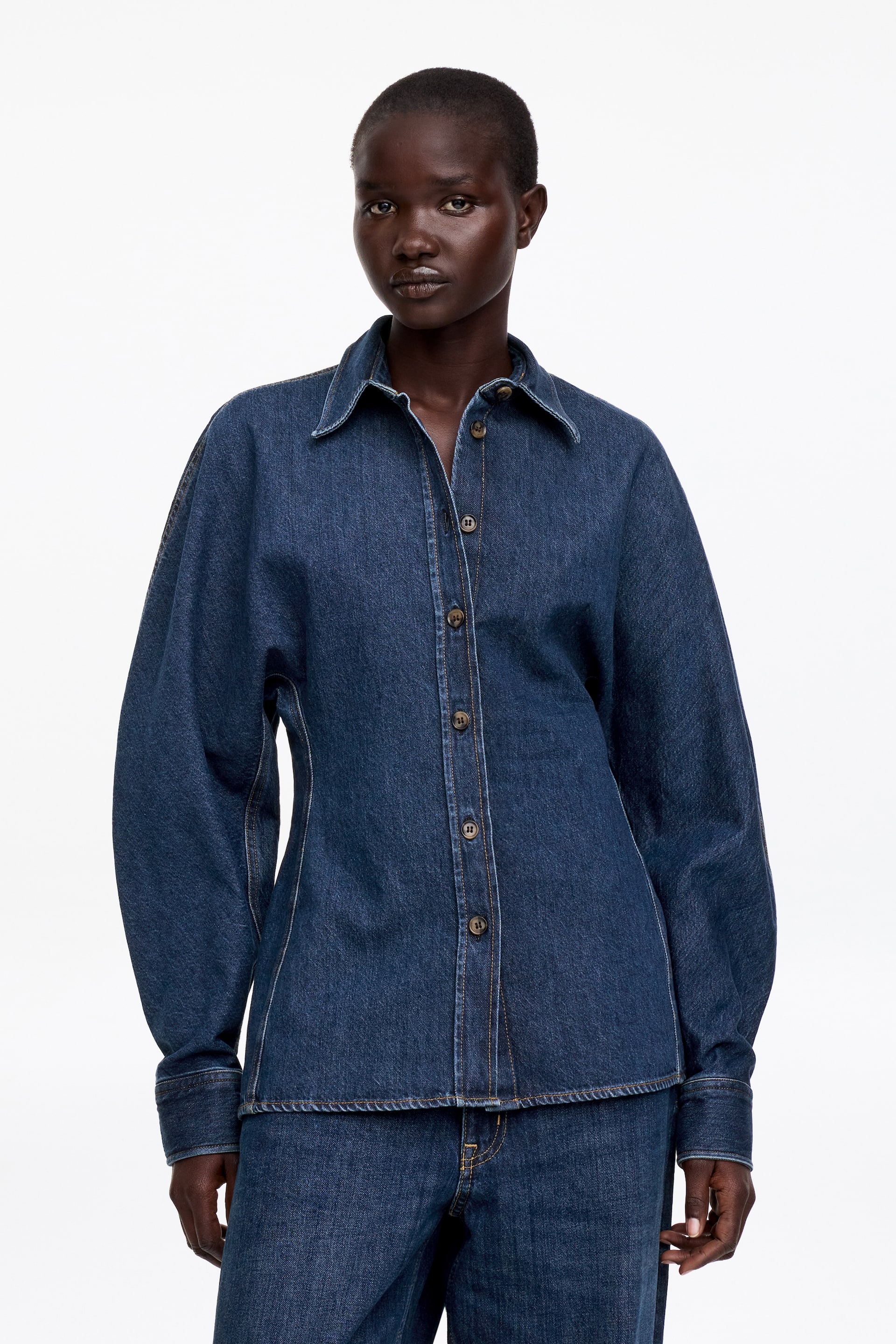 View larger image: A dark blue denim shirt featuring a pointed collar and a front button placket with light brown buttons. It is designed with long, full sleeves, buttoned cuffs, and visible light contrast stitching.
