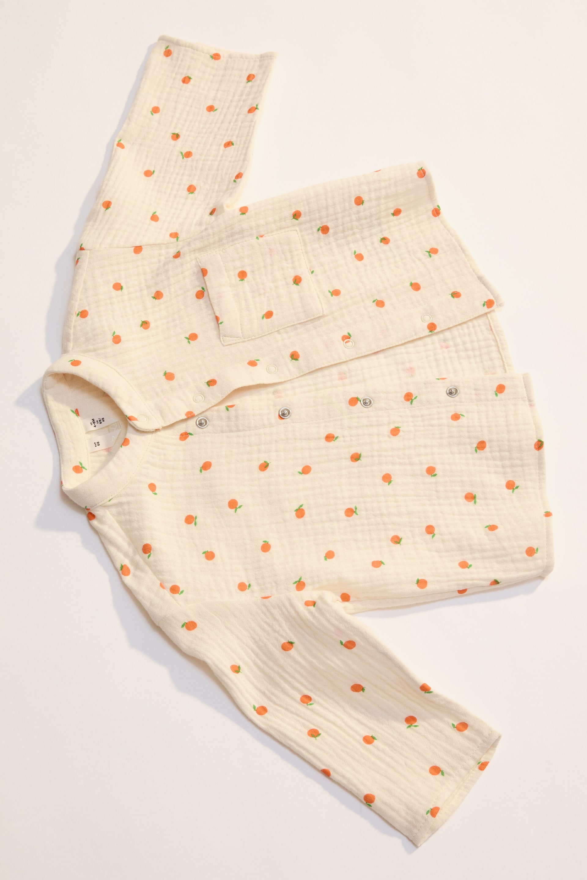 Agrandir l'image: Un body pour bébé de couleur blanc cassé est posé à plat, présentant un tissu en double gaze froissée orné de petits fruits orange. Il est conçu avec des manches longues, un col rond, une patte de boutonnage à pression s'étendant du torse en diagonale vers une jambe, et une petite poche plaquée sur la poitrine gauche.