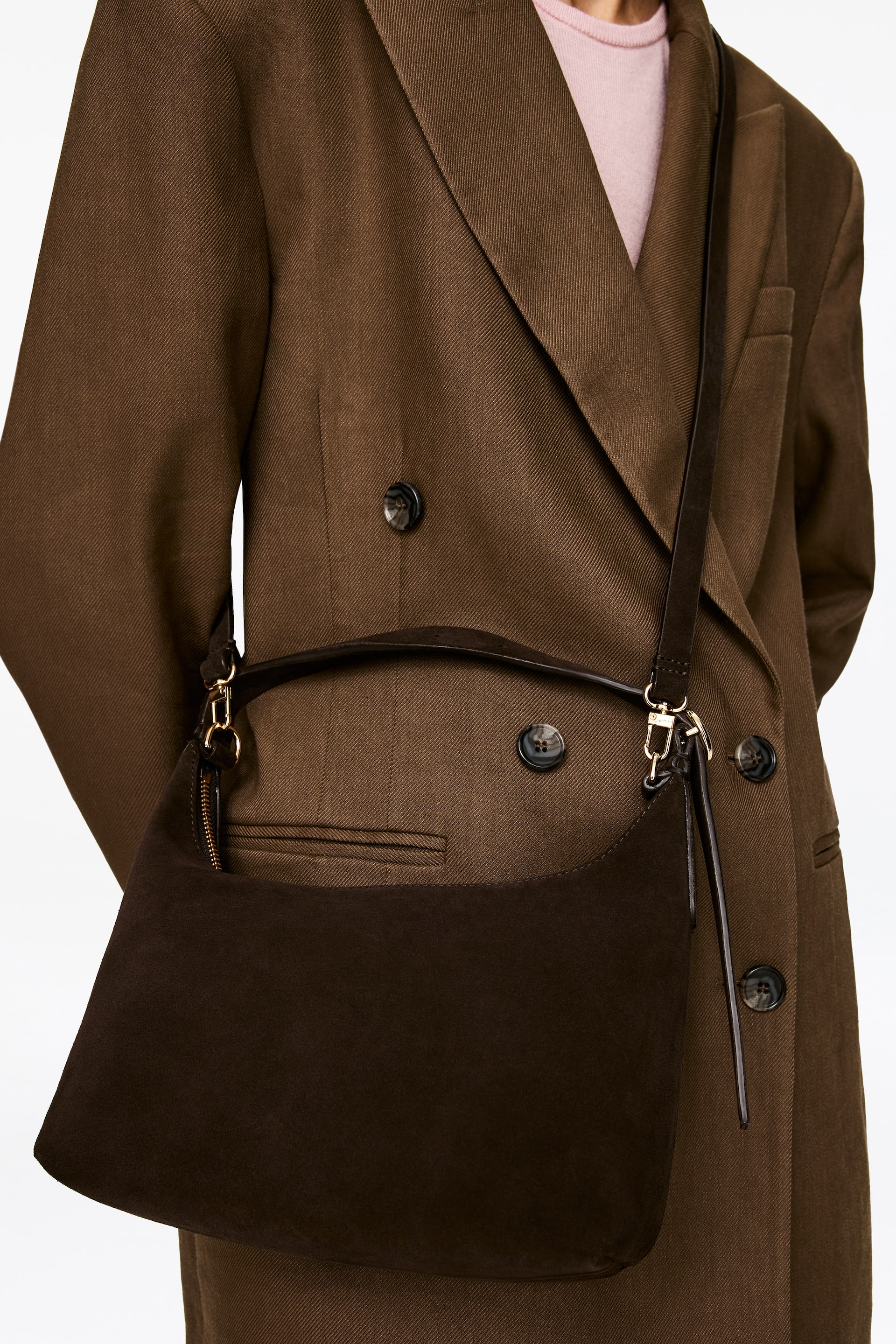 Zobraziť väčší obrázok: Tmavohnedá semišová taška visí cez telo a spočíva na hnedom dvojradovom kabáte. Má zakrivený, polmesiacový tvar so zlatým zipsovým uzáverom pozdĺž hornej hrany a je zavesená na tenkom tmavom koženom popruhu cez telo.