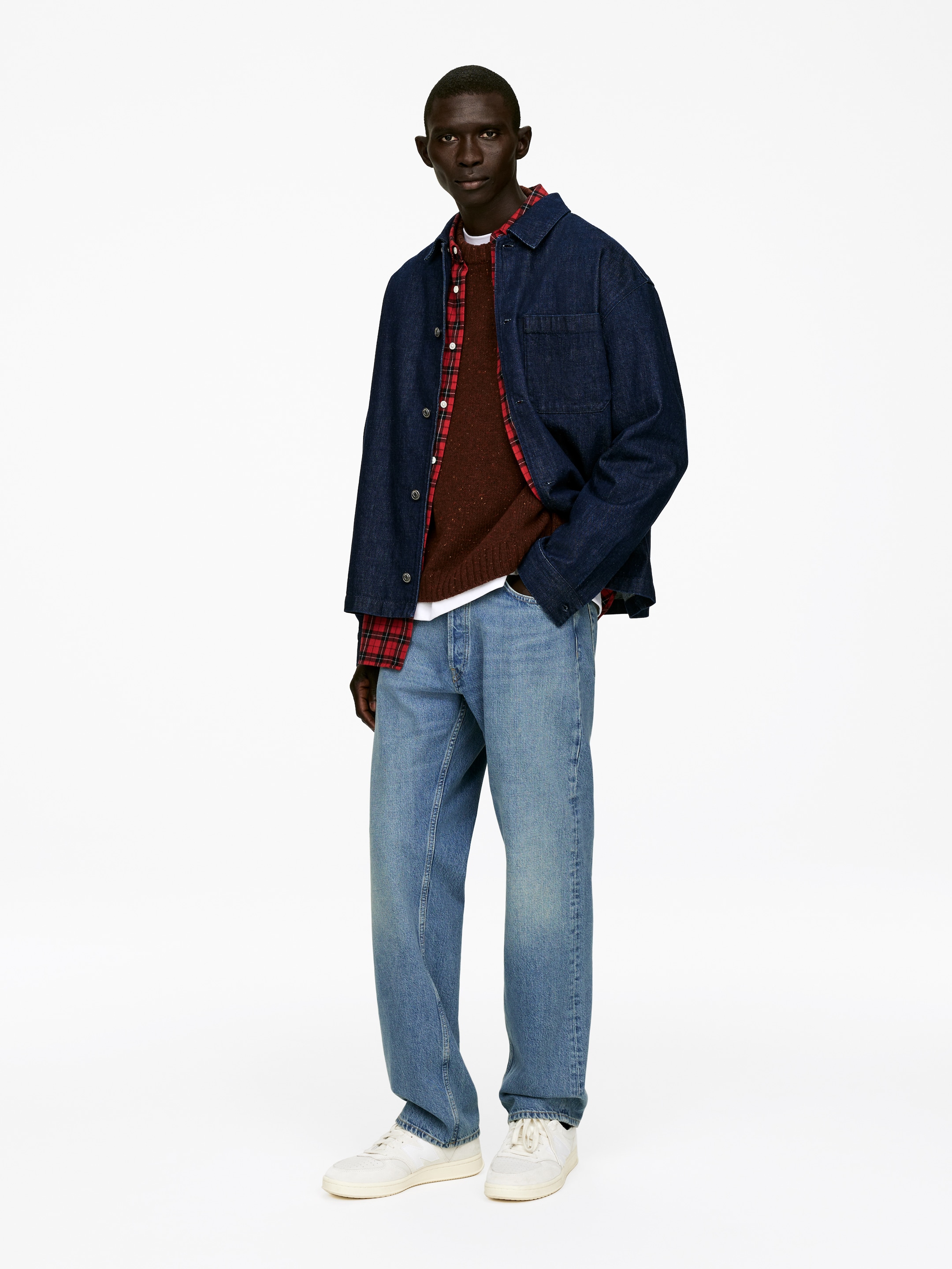 Grotere afbeelding bekijken: Een gelaagde casual outfit wordt gepresenteerd vanuit een lichte voor-rechterhoek, bestaande uit een donkerblauwe denim overshirt met een kraag en een enkele borstzak, gelaagd over een rood en zwart geruit overhemd met knopen, een bruin gespikkelde gebreide trui met ronde hals en een wit t-shirt met ronde hals. Lichtblauwe gewassen relaxed-fit jeans met rechte pijpen worden gecombineerd met witte lage vetersneakers.