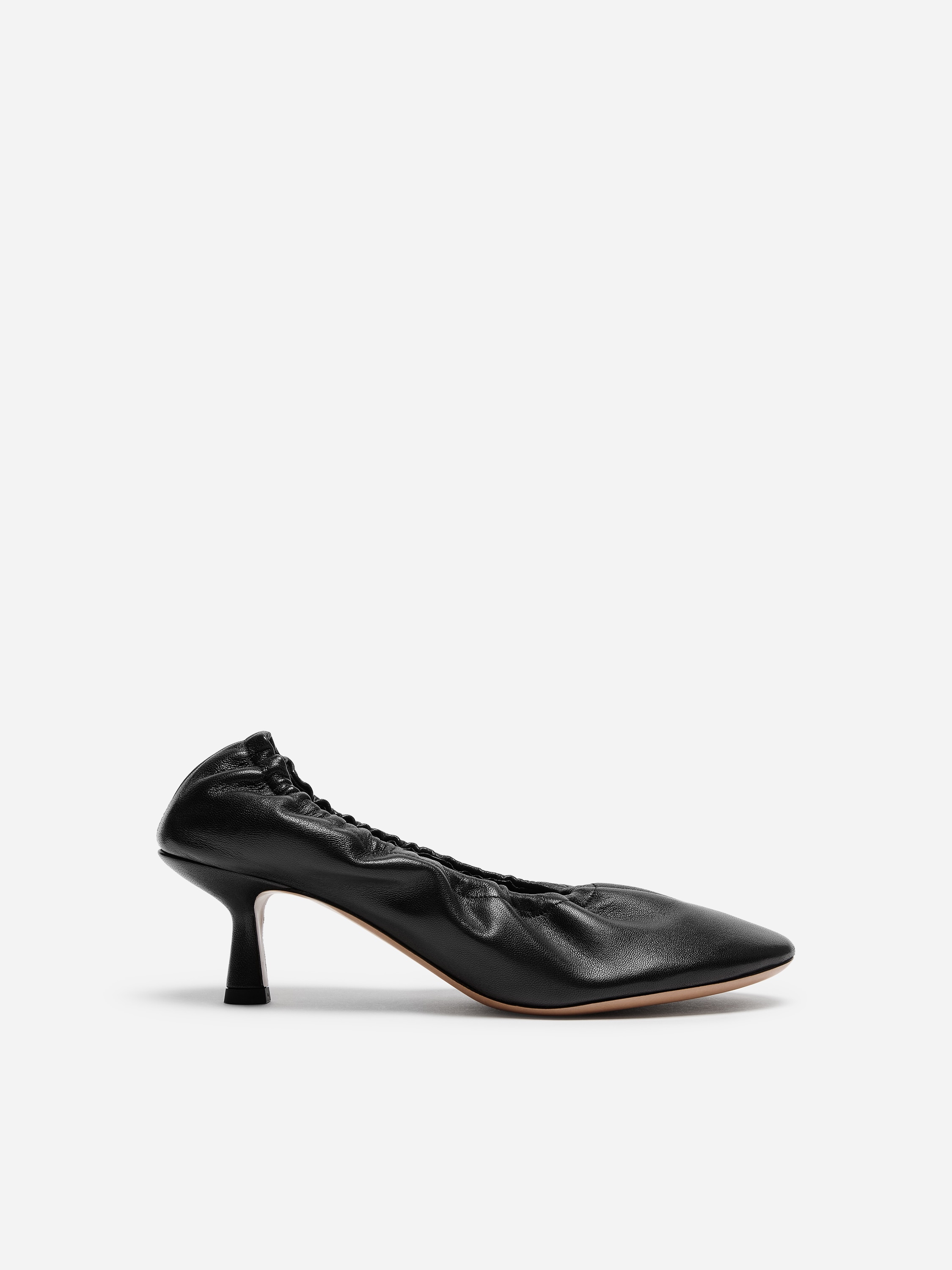 Svarte pumps i lær med avrundet tå, rynkedetaljer langs åpningen og en lav kitten heel bæres av modellen. Skoene er kombinert med svarte, transparente strømpebukser og et svart midi-skjørt.