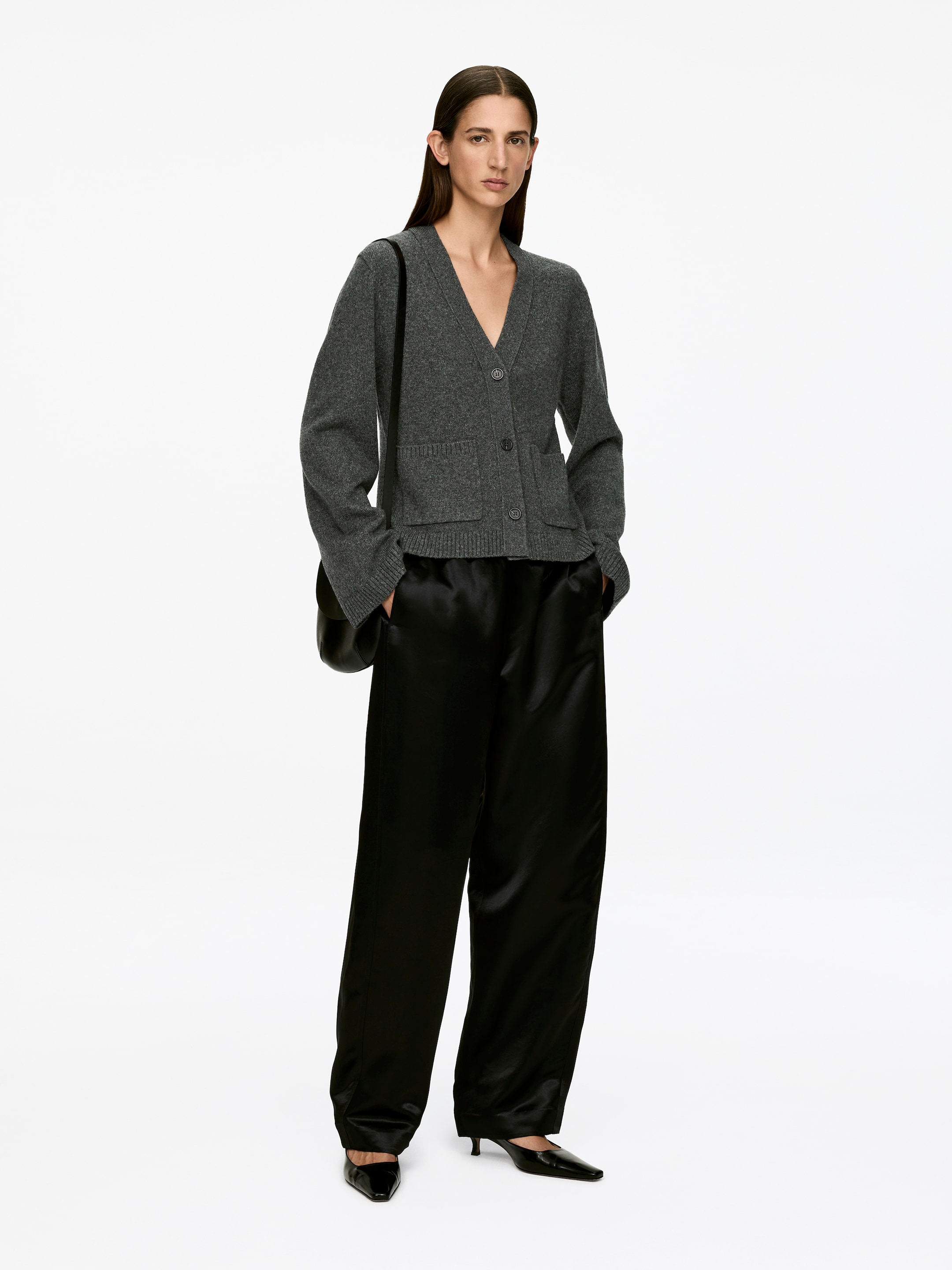 Größeres Bild ansehen: Ein grauer langärmeliger Cardigan mit V-Ausschnitt wird von dem Model getragen. Er verfügt über einen Drei-Knopf-Verschluss, der derzeit offen ist, und zwei aufgesetzte Taschen an der Vorderseite, in denen die Hände des Models ruhen. Der Cardigan wird über schwarze Hosen gestylt.