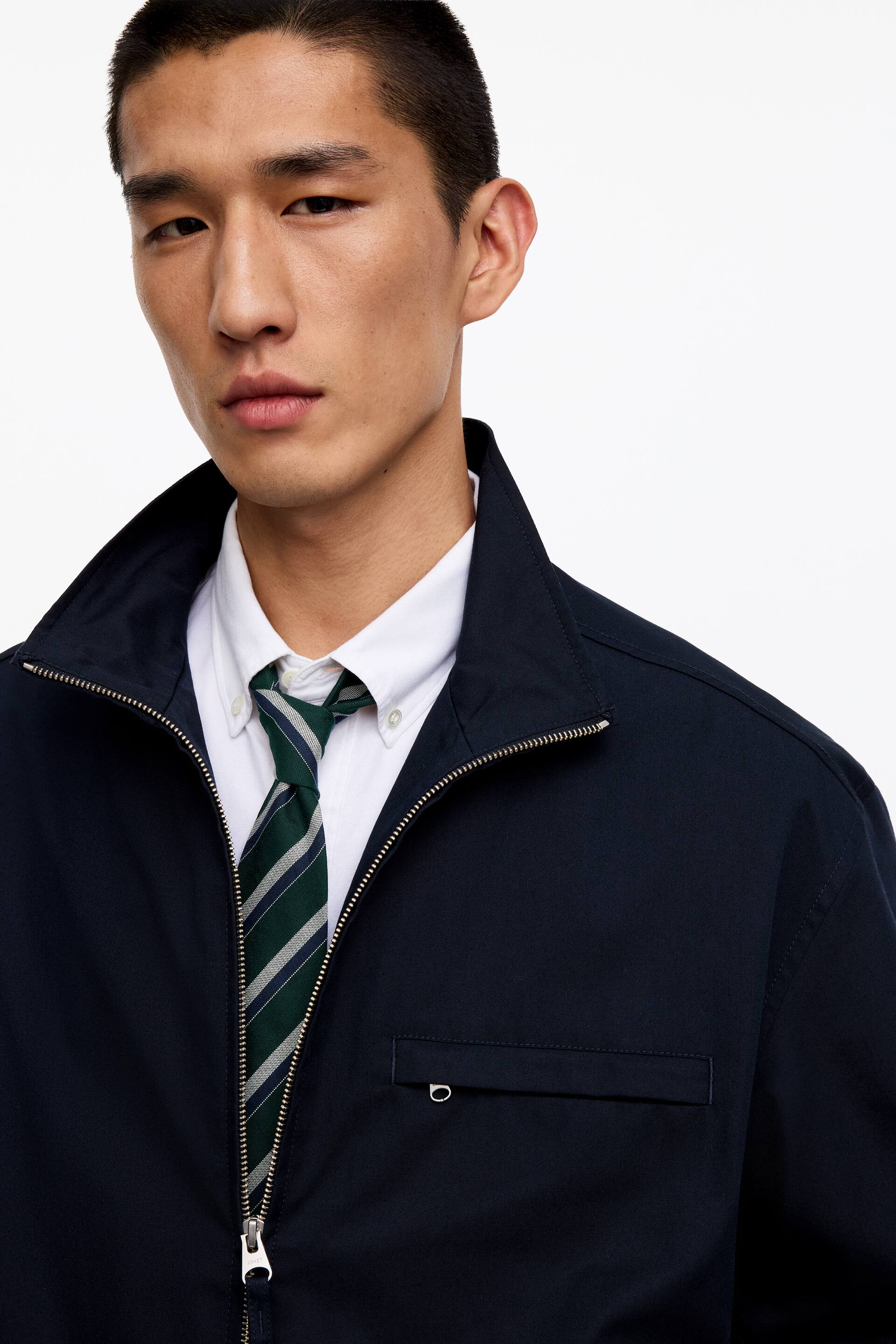 Ver imagem maior: Uma gravata de seda, com riscas diagonais verdes, azul-escuro e brancas, está a ser usada pelo modelo. Está atada à gola de uma camisa branca, visível por baixo de um casaco azul-escuro parcialmente aberto.