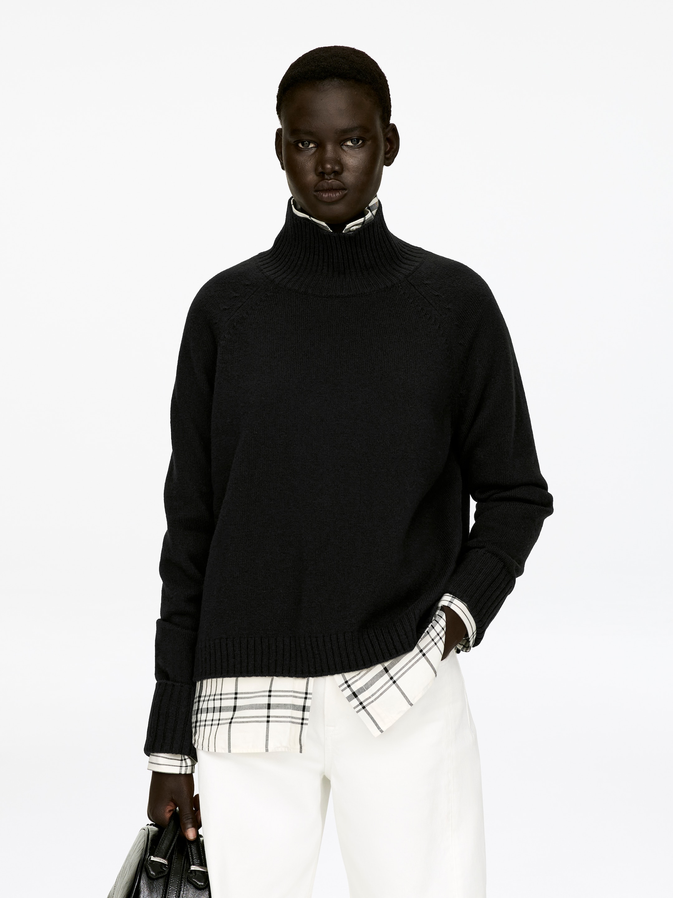 View larger image: A black roll-neck jumper, featuring raglan sleeves and folded cuffs, is being worn by the model over a white and black plaid shirt, which is visible at the neck and extending below the jumper's hem. The model is also wearing white trousers and holding a black bag.