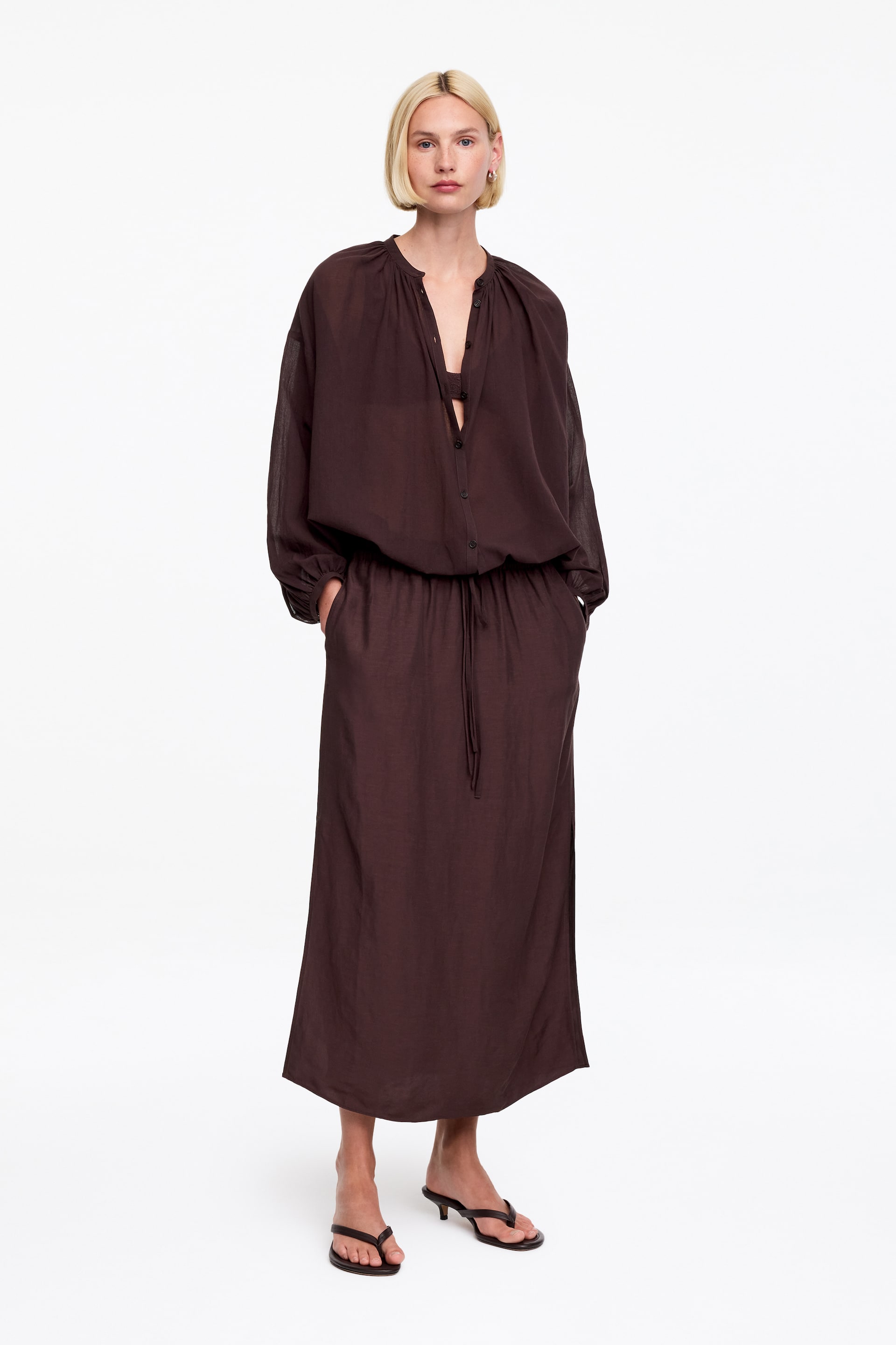 Visualizza immagine più grande: Un abito maxi marrone scuro presenta un top blouson trasparente con un'allacciatura parziale a bottoni e uno scollo arricciato, insieme a maniche lunghe e voluminose che terminano con polsini elasticizzati. Un reggiseno marrone scuro è visibile sotto il top trasparente, che scende in una gonna lunga fino ai piedi, stretta in vita da un cordoncino.