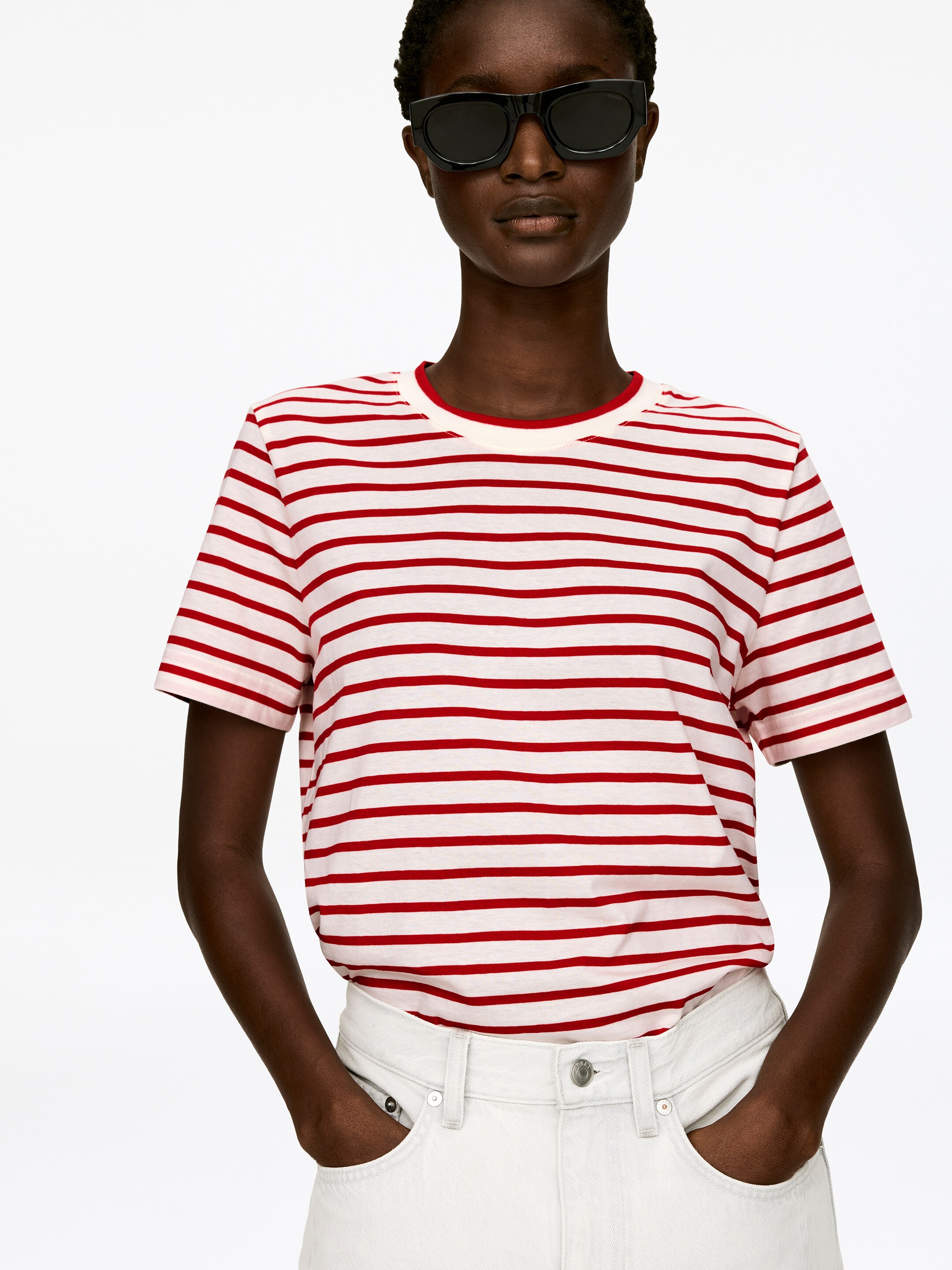Grösseres Bild ansehen: Ein T-Shirt wird von vorne präsentiert und zeigt horizontale rote und cremefarbene Streifen. Es hat einen runden Halsausschnitt mit rotem Besatz und kurze Ärmel und ist in eine hellfarbige Hose gesteckt.