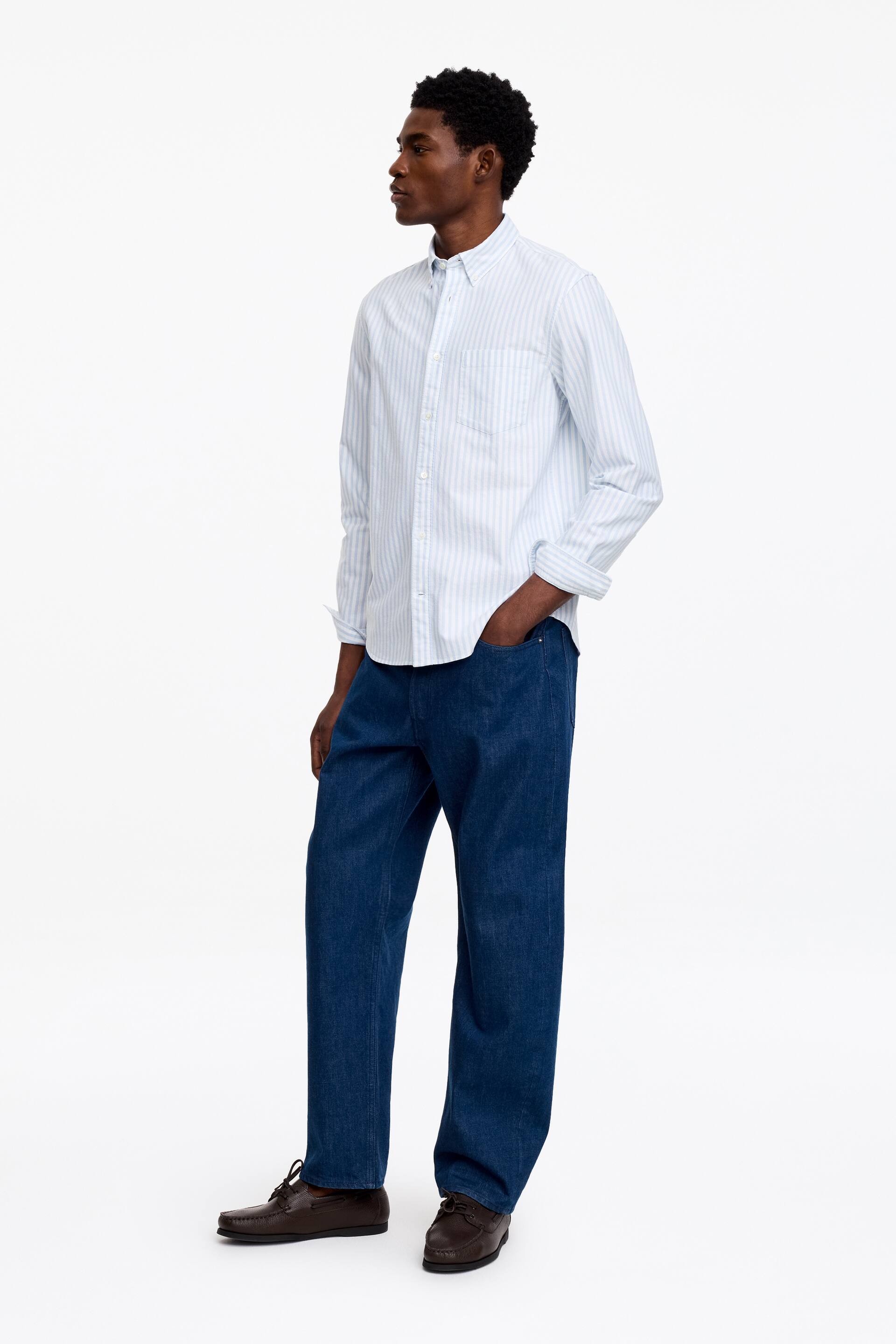 Agrandir l'image: Une chemise à manches longues à rayures verticales bleu clair et blanc est présentée sous un léger angle latéral, montrant un col à boutons, une poche poitrine plaquée et des manches retroussées jusqu'aux avant-bras.