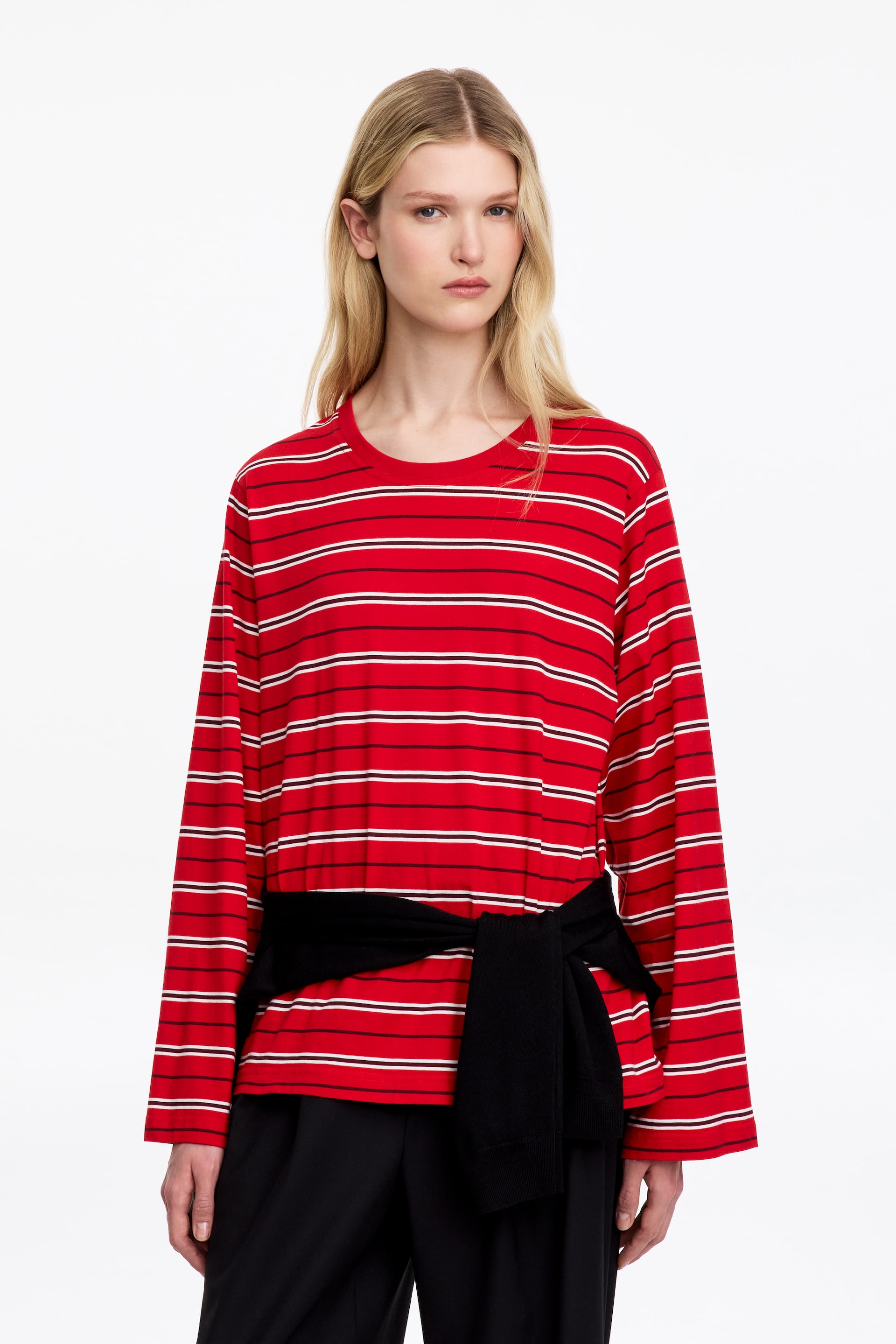 Ver imagem maior: O modelo está a usar uma T-shirt vermelha de manga comprida, com riscas horizontais pretas e brancas. A peça de gola redonda, de corte largo, é combinada com uma camisola preta atada à cintura e umas calças pretas.