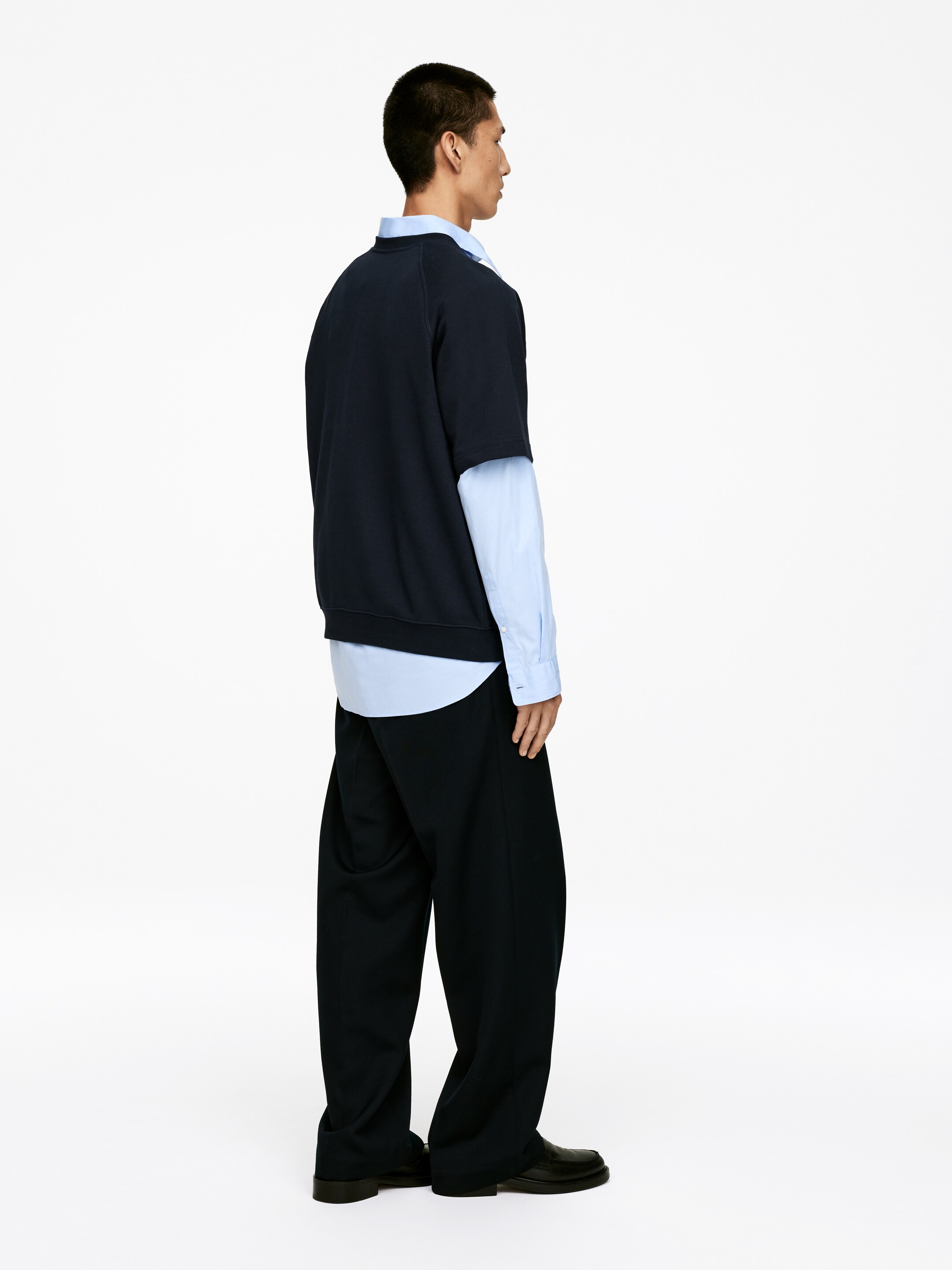 Ingrandisci l'immagine: Una felpa a maniche corte blu scuro, con maniche raglan e orlo a coste, è indossata a strati sopra una camicia azzurra a maniche lunghe con colletto. Pantaloni a gamba larga blu scuro e scarpe nere lucide sono anch'essi indossati, presentando un profilo laterale completo dell'outfit a strati.