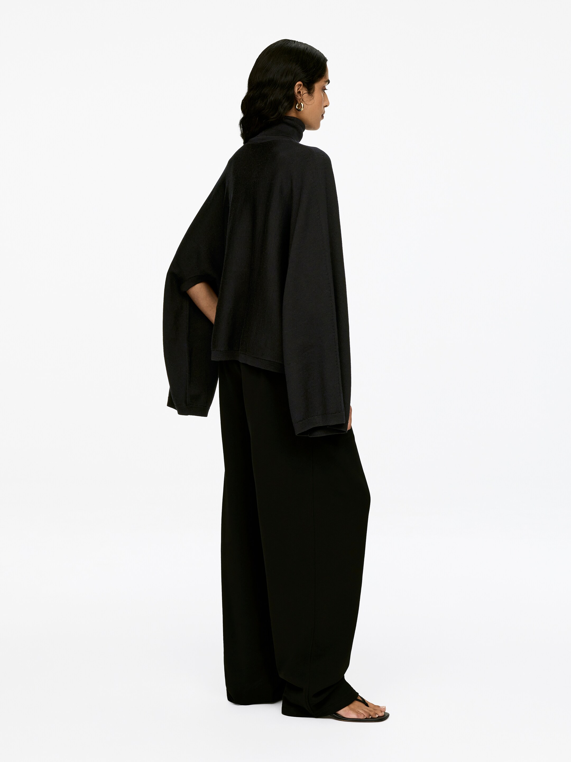 Agrandir l'image: Un pull à col roulé noir avec une coupe ample est porté par le mannequin. Un des bras du mannequin s'étend à travers une ouverture visible, tandis que l'autre bras reste dans la silhouette drapée du pull. Le pull est assorti de pantalons noirs à jambes larges.