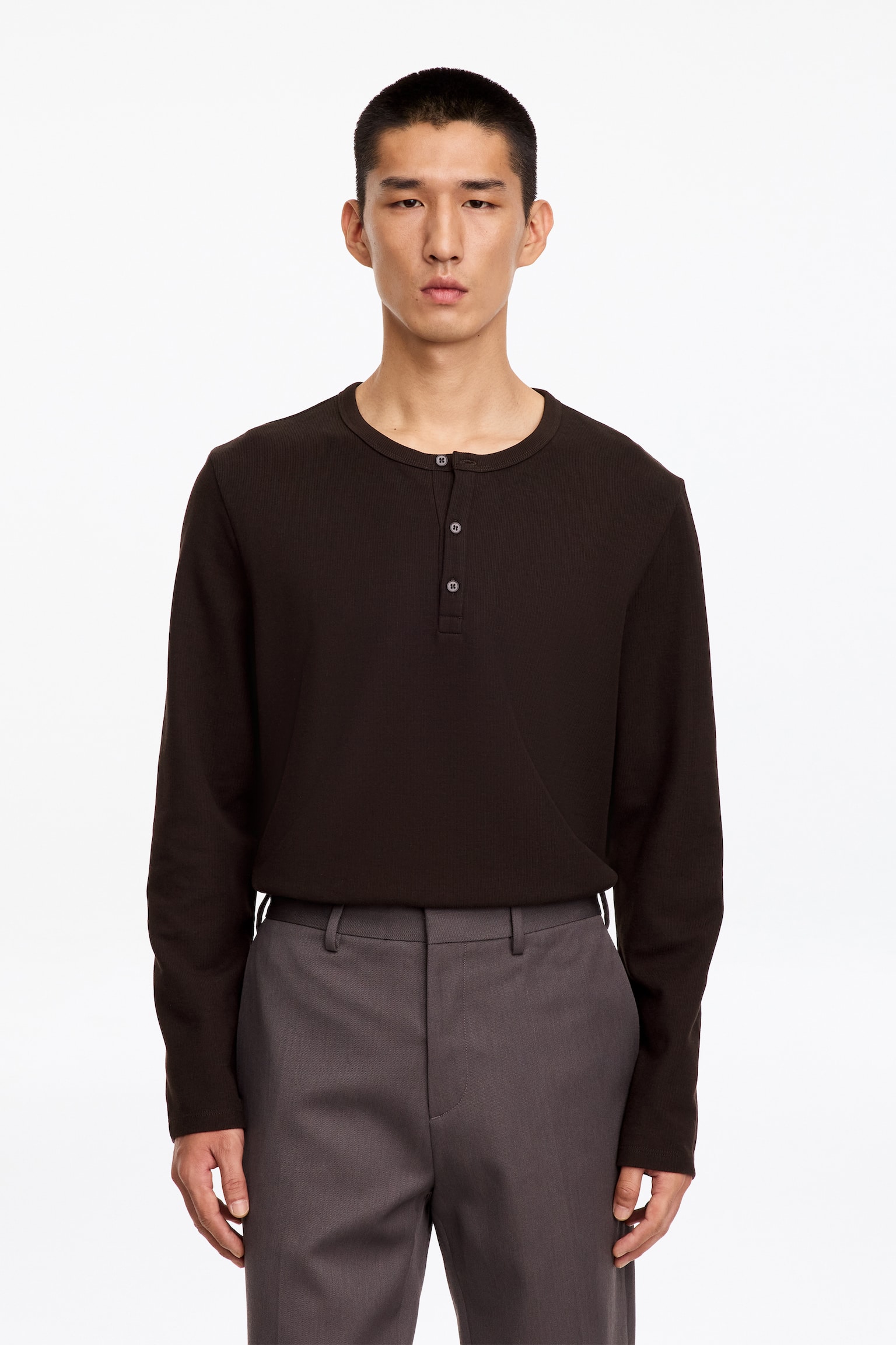 Un haut Henley à manches longues brun foncé, en maille côtelée, est porté par le mannequin. Le haut présente un col rond côtelé et une patte de boutonnage à demi-ouverture, et il est rentré dans un pantalon gris tailleur. - 1