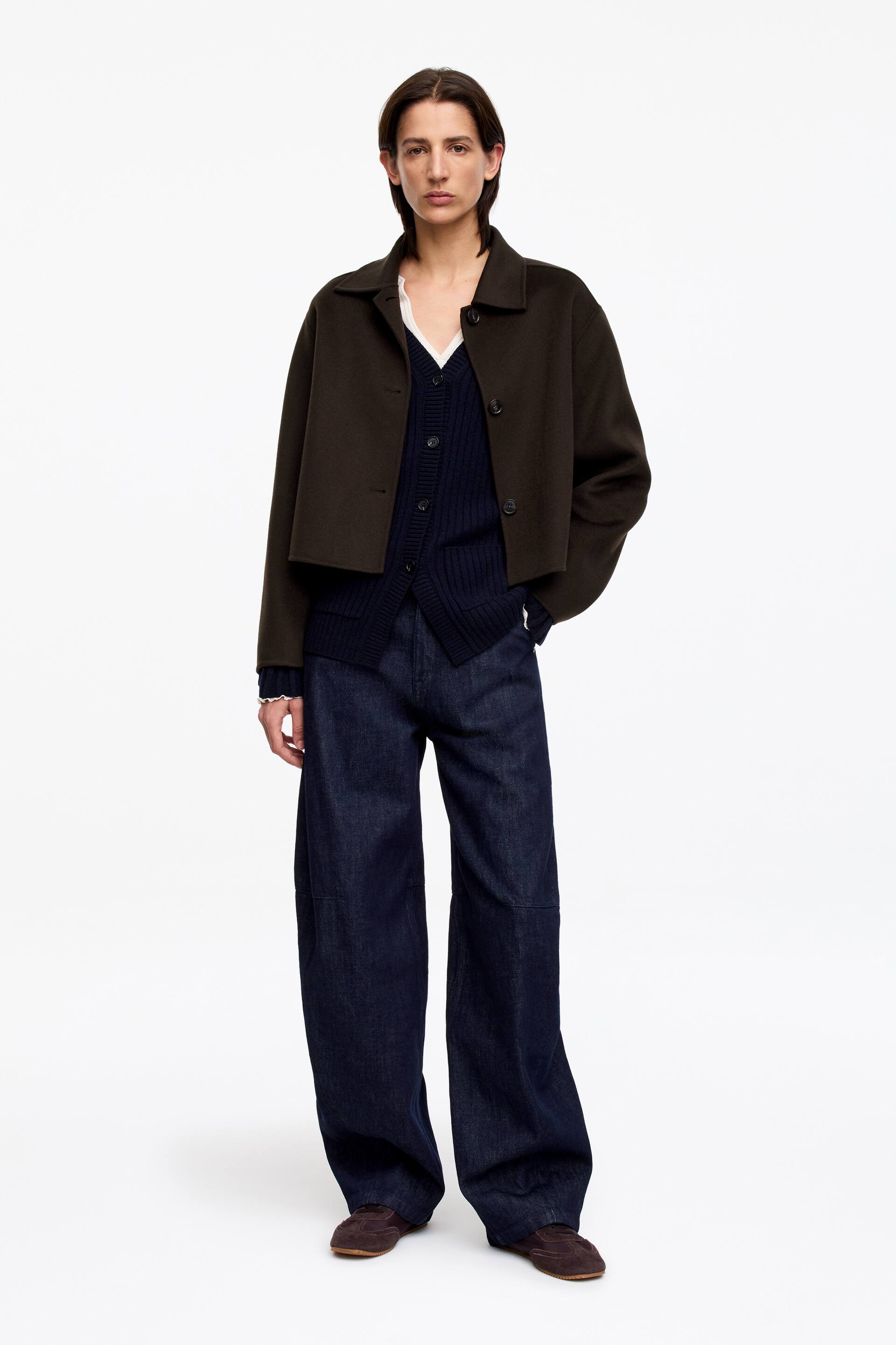 Größeres Bild anzeigen: Eine dunkelolivfarbene, kurze Jacke mit Kragen und sichtbaren Frontknöpfen wird über einem marineblauen, gerippten V-Ausschnitt-Cardigan getragen, der offen über einer weißen Bluse mit Rüschenmanschetten liegt. Dunkelblaue, weit geschnittene Jeanshosen vervollständigen das nach vorne gerichtete Ensemble, getragen mit braunen flachen Schuhen.