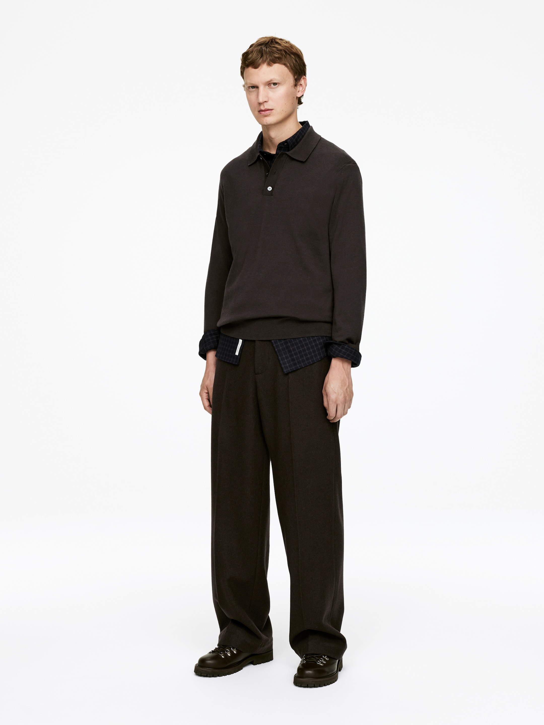 Agrandir l'image: Un polo à manches longues en tricot marron foncé, avec un col et un bouton blanc visible, est porté en superposition sur une chemise à carreaux foncés dont le col et les manches retroussées sont visibles. Cela est assorti à un pantalon large marron foncé qui s'accumule sur des bottes épaisses et sombres avec des œillets en métal, l'ensemble étant présenté sous un léger angle frontal.