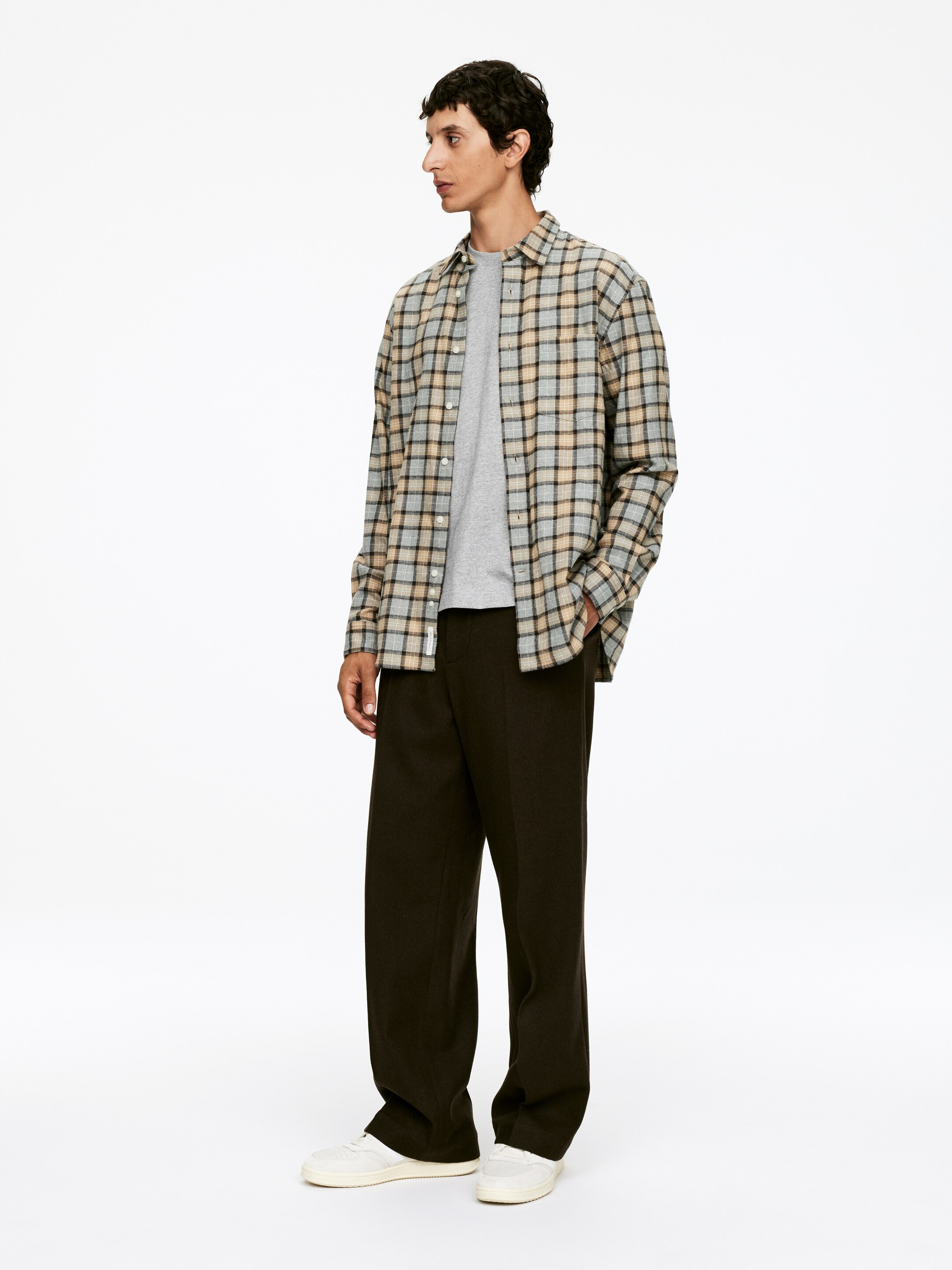 Visa större bild: En modell står i en lätt vinkel och visar upp en öppen rutig skjorta med ljusblå, beige och svarta rutor, som är lagd över en enkel grå t-shirt. Looken kompletteras med vida mörkbruna byxor som har ett avslappnat fall och vita låga sneakers.