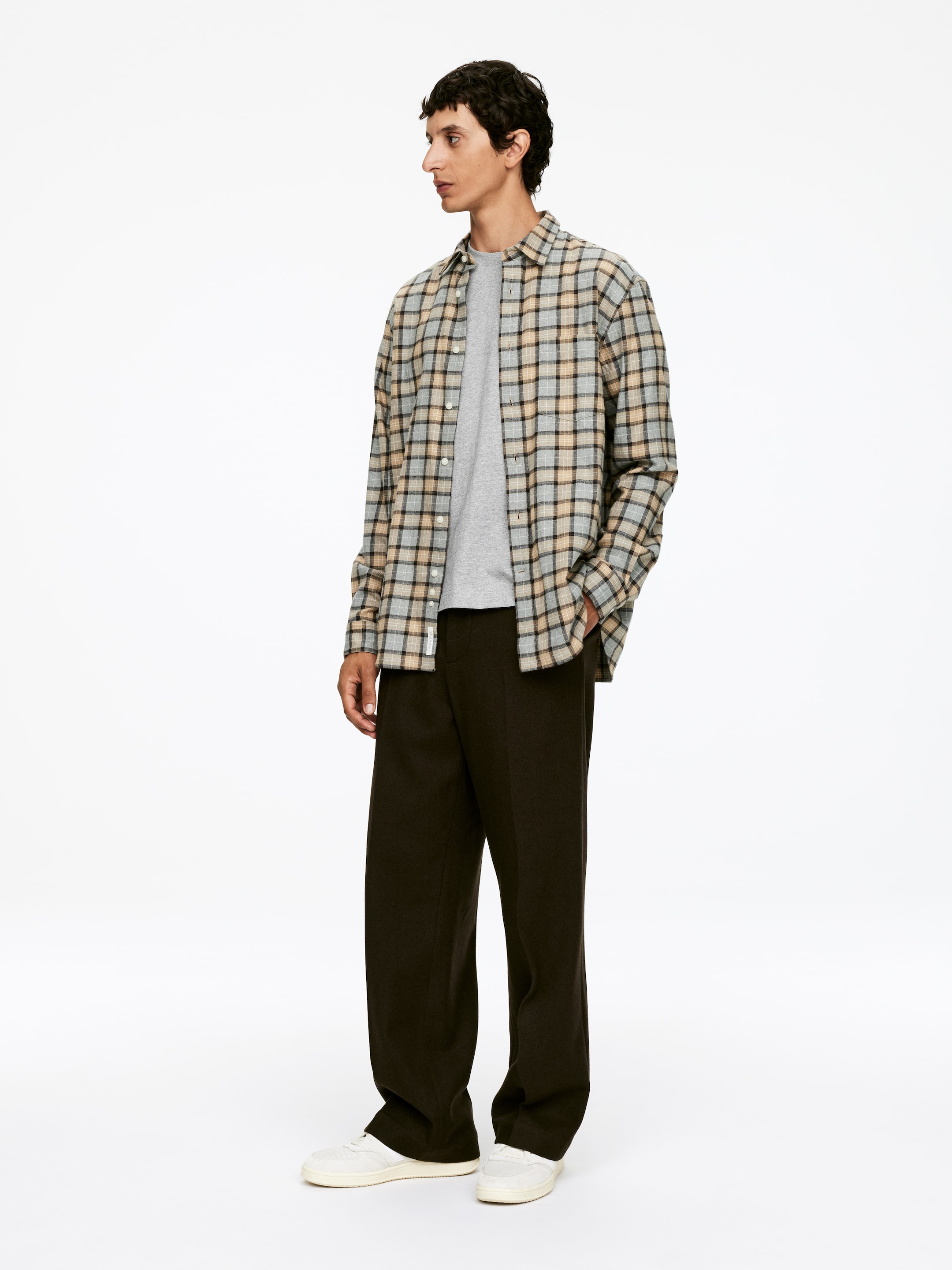 Visualizza immagine più grande: Una modella è in piedi con un leggero angolo, mostrando una camicia a quadri aperta con quadretti azzurri, beige e neri, che è stratificata su una t-shirt grigia semplice. Il look è completato da pantaloni marrone scuro a gamba larga, che presentano una caduta rilassata, e sneakers bianche a collo basso.