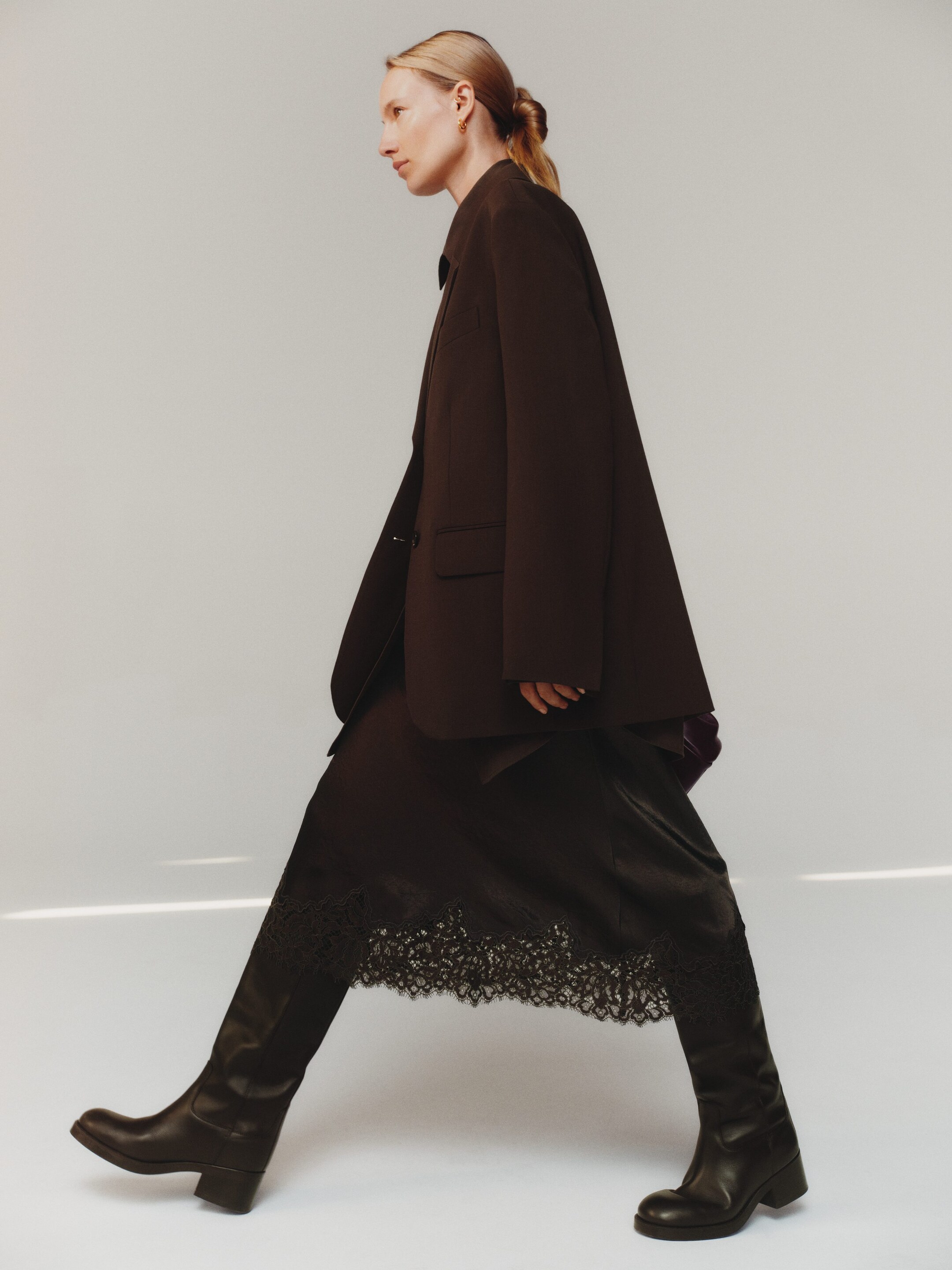 Visualizza immagine più grande: Viene presentato da una prospettiva laterale un blazer oversize marrone scuro con revers a lancia, indossato sopra una gonna midi scura con un elaborato bordo in pizzo lungo l'orlo, e abbinato a stivali scuri alti fino al ginocchio con tacco a blocco.