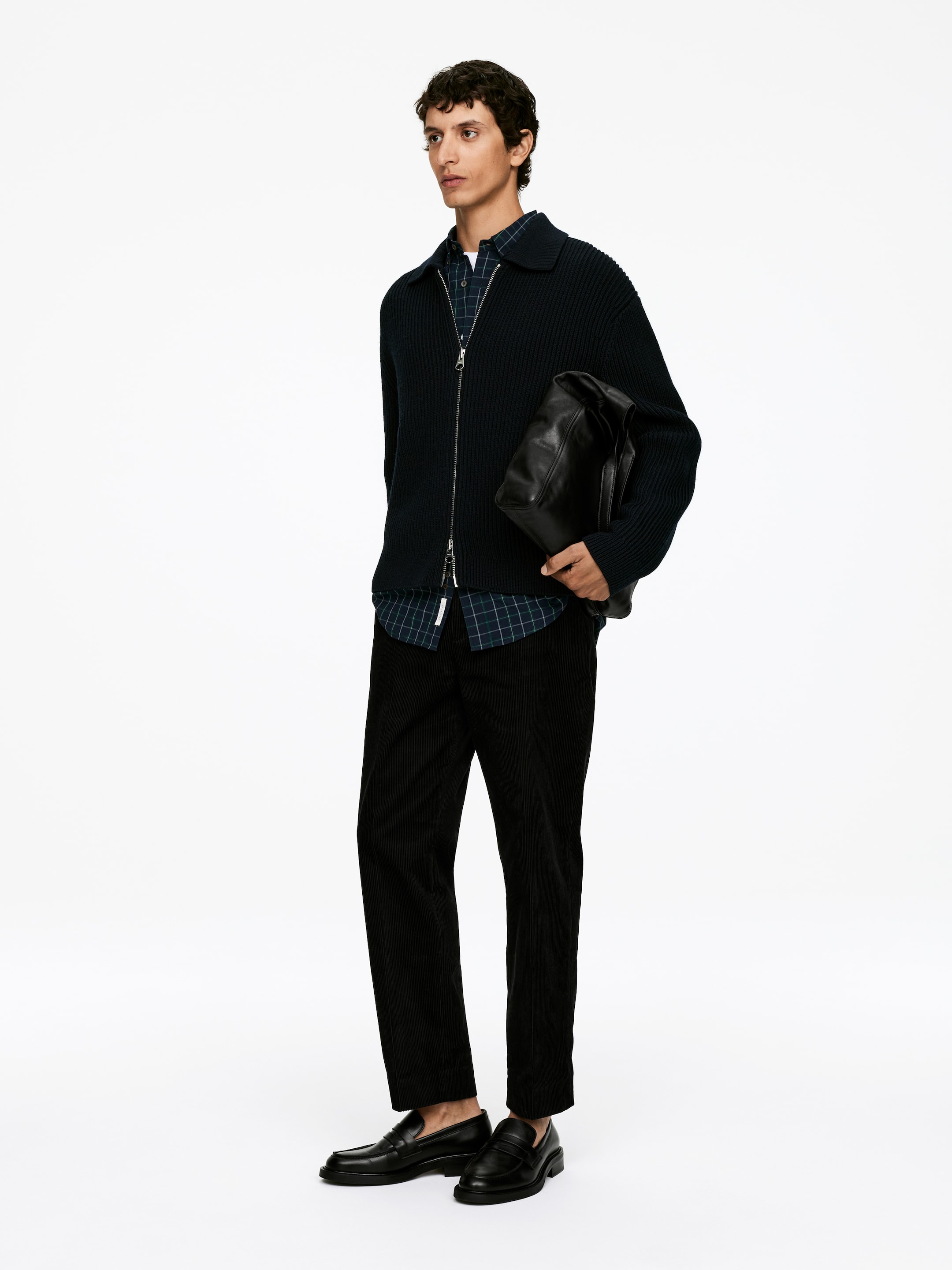 Visa större bild: En outfit har en mörk marinblå ribbstickad kofta med full dragkedja, bärs öppen över en rutig långärmad skjorta. Ensemblen kompletteras med svarta manchesterbyxor, svarta läderloafers och en svart läderväska som hålls i handen.