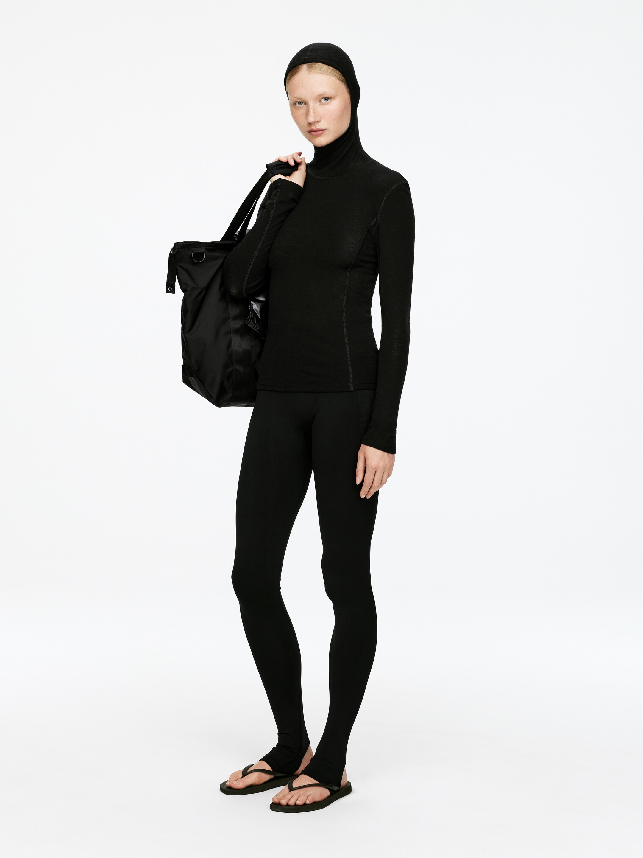 Visualizza immagine più grande: Un top nero aderente a maniche lunghe con collo alto, con dettagli di cuciture visibili e un motivo testurizzato sottile, è indossato con un passamontagna nero abbinato che copre la testa. Leggings neri aderenti con staffe si estendono su infradito nere, mentre una borsa a tracolla nera è portata sulla spalla destra.
