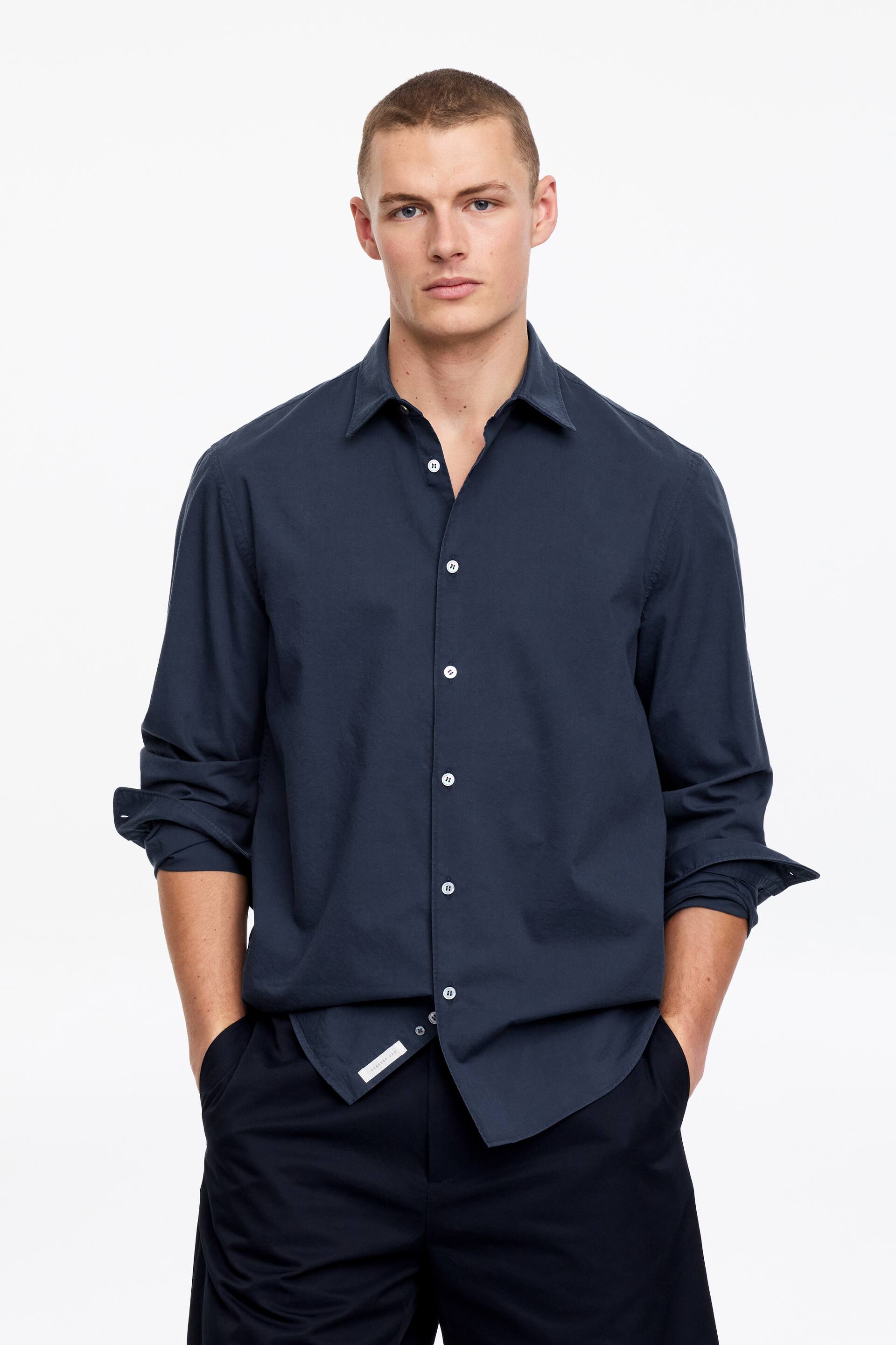 Grotere afbeelding bekijken: Een donkerblauw overhemd met lange mouwen wordt getoond, voorzien van een spreadkraag en een knoopsluiting met witte knopen. De mouwen van het overhemd zijn opgerold tot aan de onderarmen en een klein wit label is zichtbaar nabij de linker onderzoom.