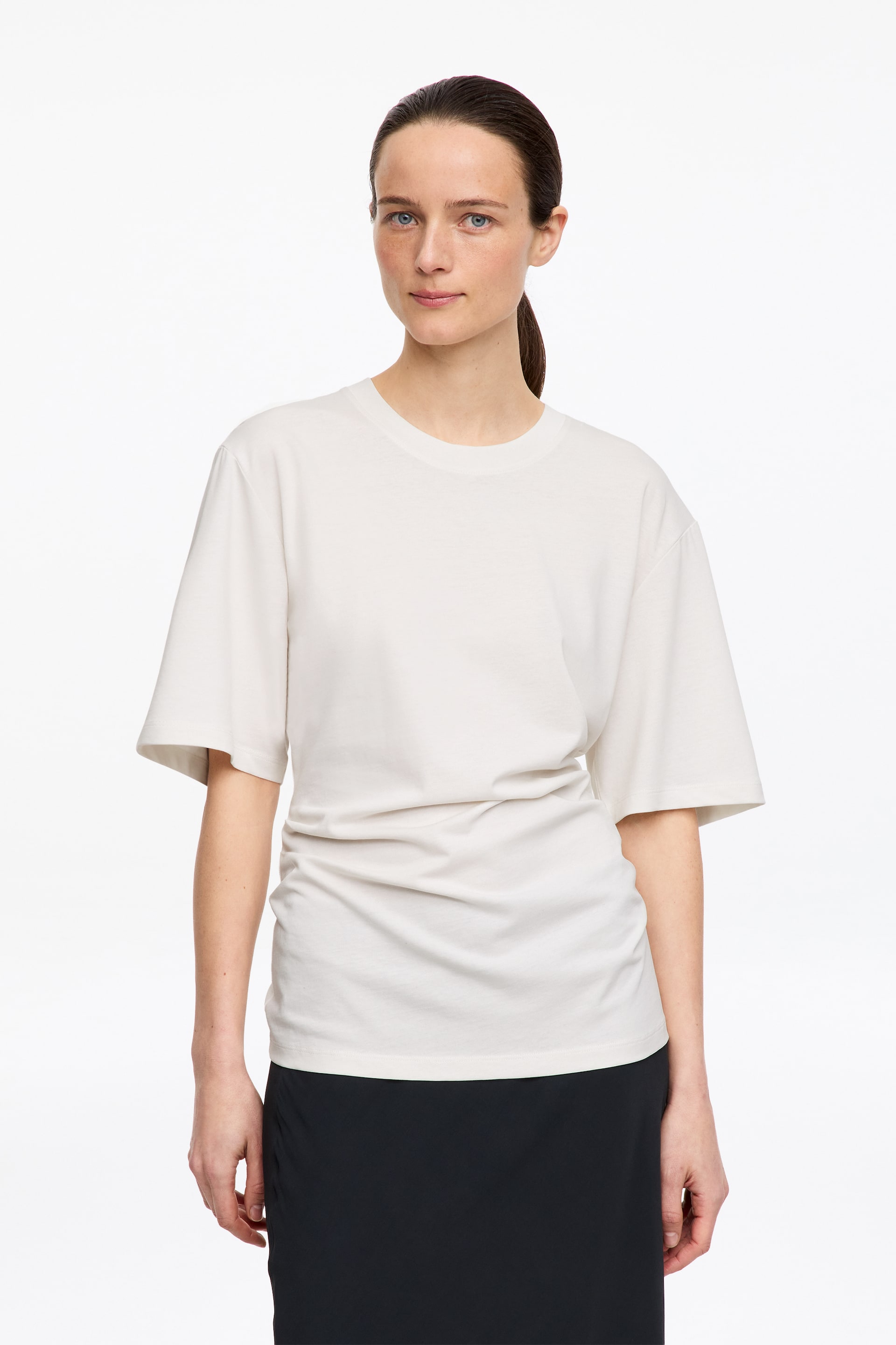 Grotere afbeelding bekijken: Een off-white t-shirt met ronde hals is naar voren gericht en heeft een ontspannen pasvorm met brede, korte mouwen. Het t-shirt toont plooidetails langs de linkerkant van de taille, wat een gedrapeerd silhouet creëert wanneer het over een donkere rok wordt gedragen.
