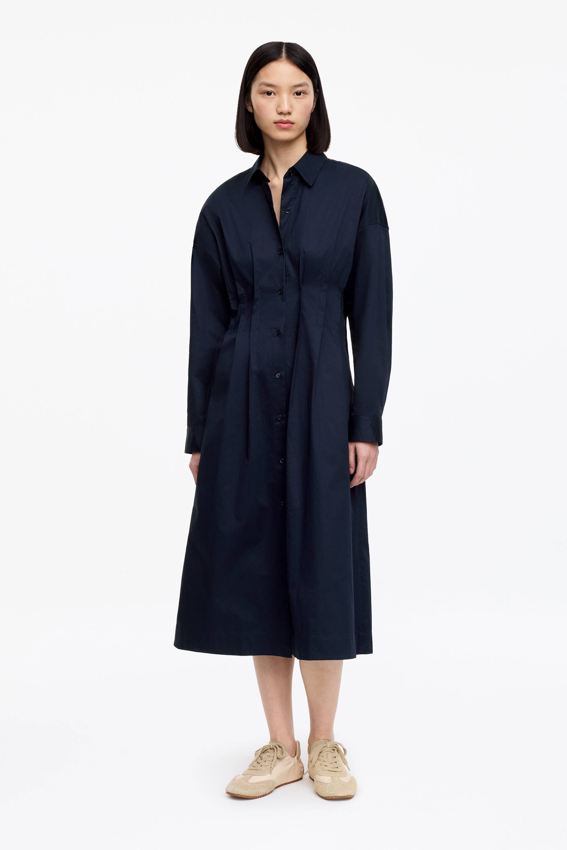 Größeres Bild ansehen: Ein marineblaues Hemdblusenkleid präsentiert einen klassischen spitzen Kragen und lange Ärmel mit geknöpften Manschetten. Es verfügt über eine durchgehende Knopfleiste, eine taillierte Taille mit vertikalen Falten und einen A-Linien-Midirock.