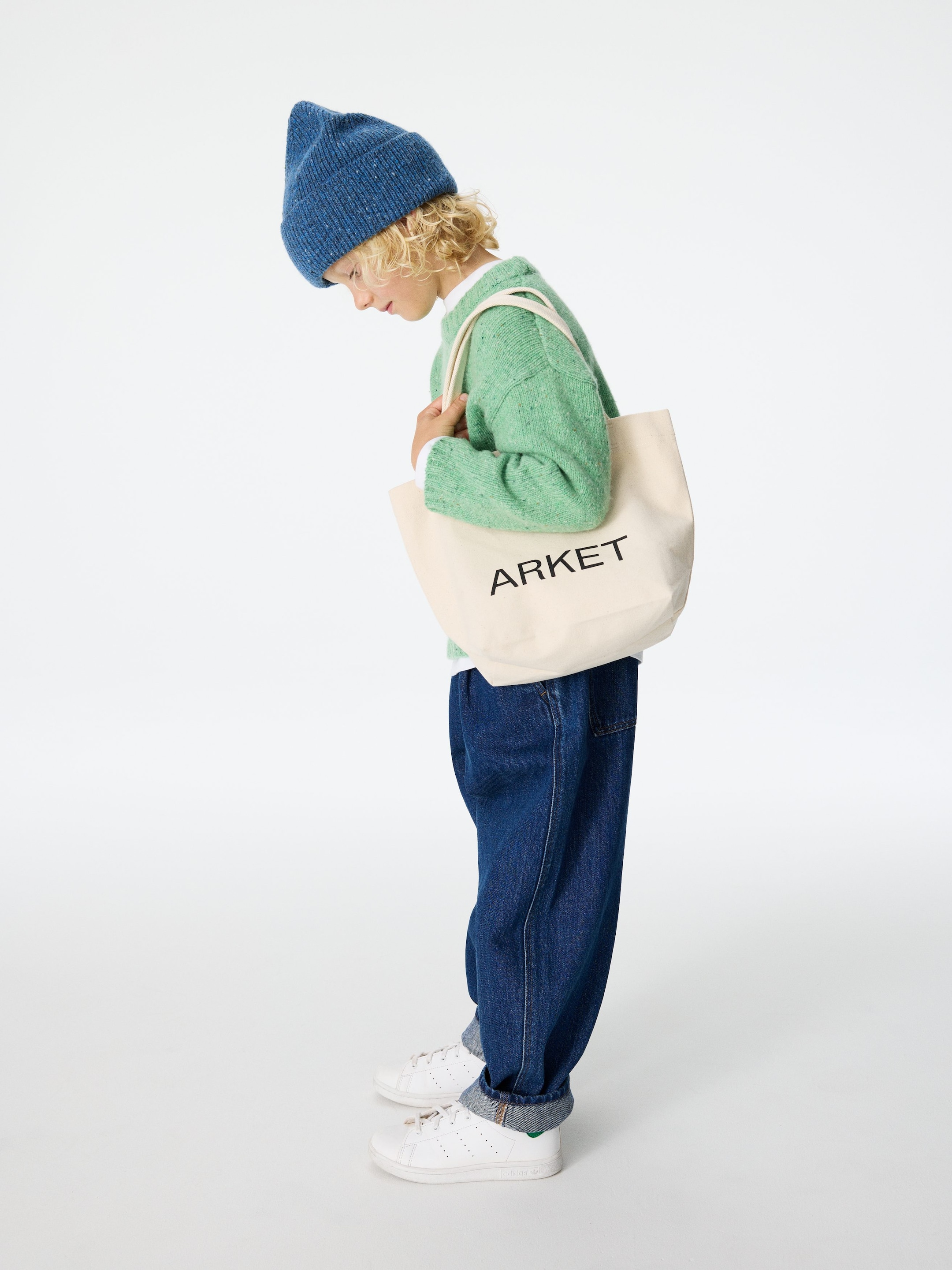 Größeres Bild anzeigen: Das Produkt, präsentiert in einer Seitenansicht, zeigt eine blaue gestrickte Mütze mit gesprenkeltem Muster und umgeschlagenem Rand, einen hellgrünen gestrickten Pullover mit gesprenkeltem Muster und gerippten Bündchen, der über einem weißen Hemd getragen wird, und dunkelblaue Jeans mit weitem Bein und umgeschlagenen Säumen. Weiße Low-Top-Sneaker vervollständigen das Ensemble, und eine cremefarbene Stofftasche mit 'ARKET'-Branding wird vorne gehalten.