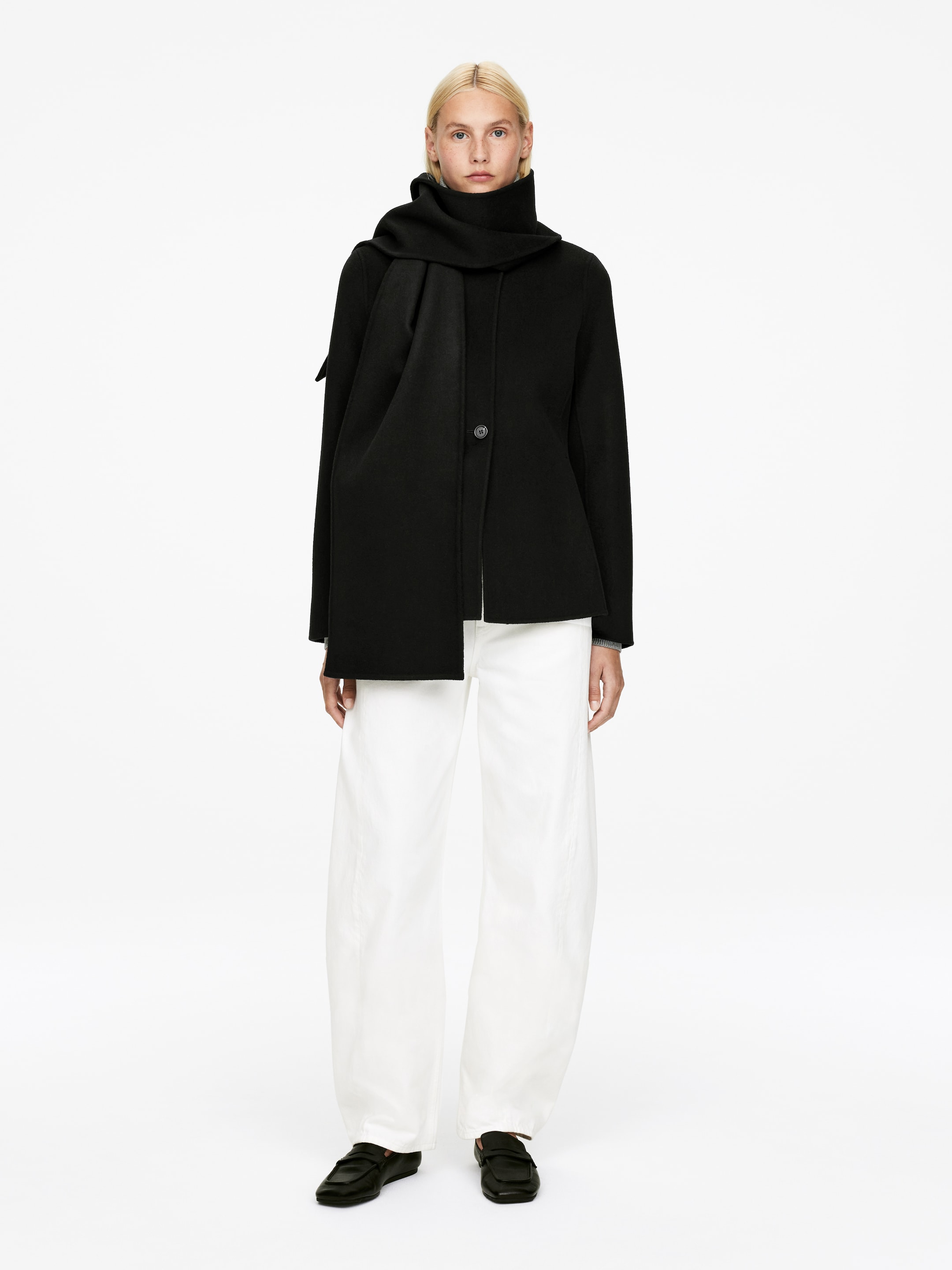 Agrandir l'image: Un manteau court noir est présenté de face, avec une fermeture à un bouton et une écharpe noire drapée autour du cou et sur le côté gauche. Le manteau est superposé à un pantalon large blanc, assorti à des mocassins plats noirs.