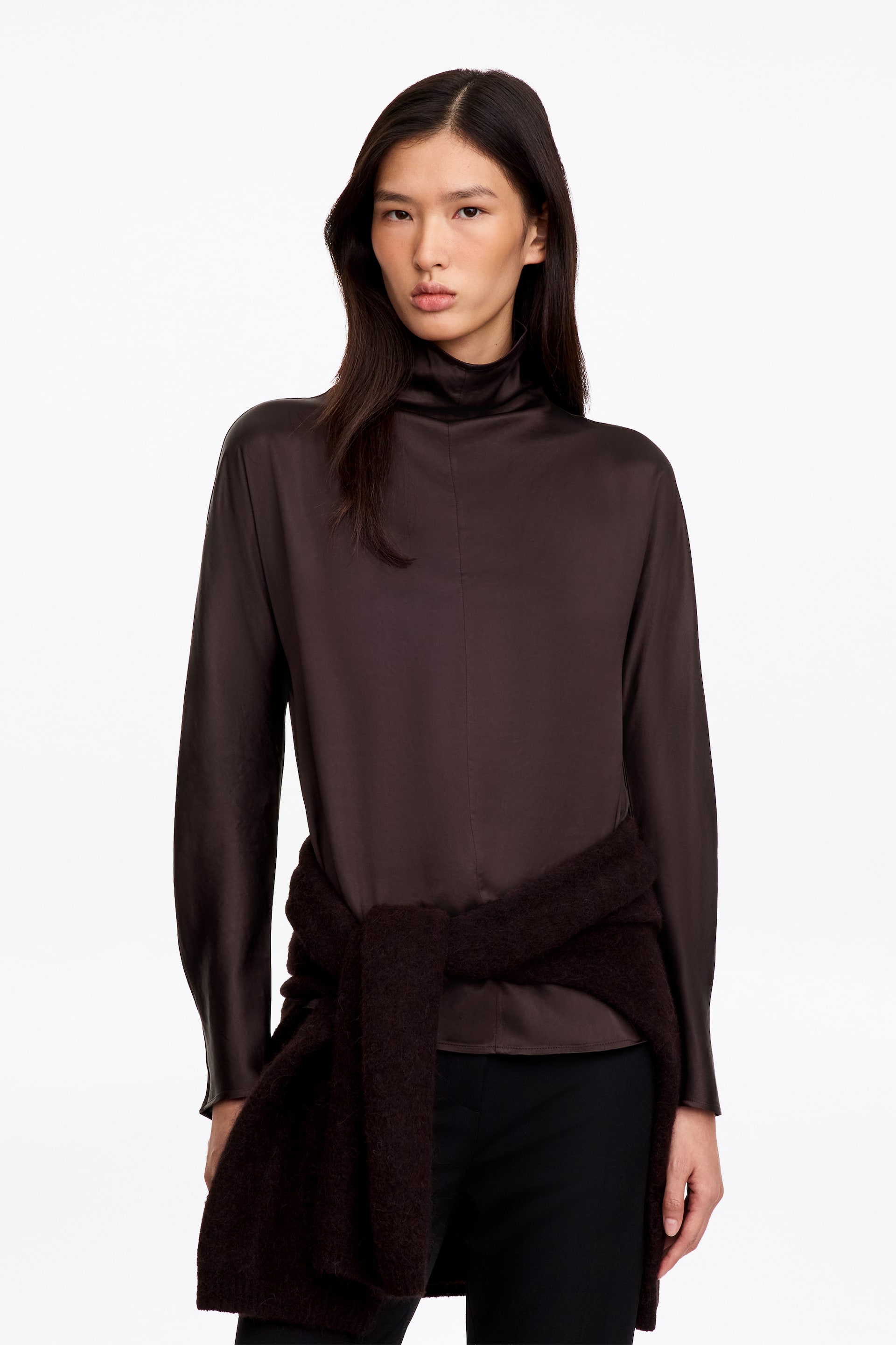 Visualizza immagine più grande: La modella indossa un top a collo alto finto in raso marrone scuro, con maniche lunghe e cucitura centrale sul davanti. Il top è abbinato a pantaloni neri e a un maglione lavorato a maglia scuro e testurizzato annodato in vita.
