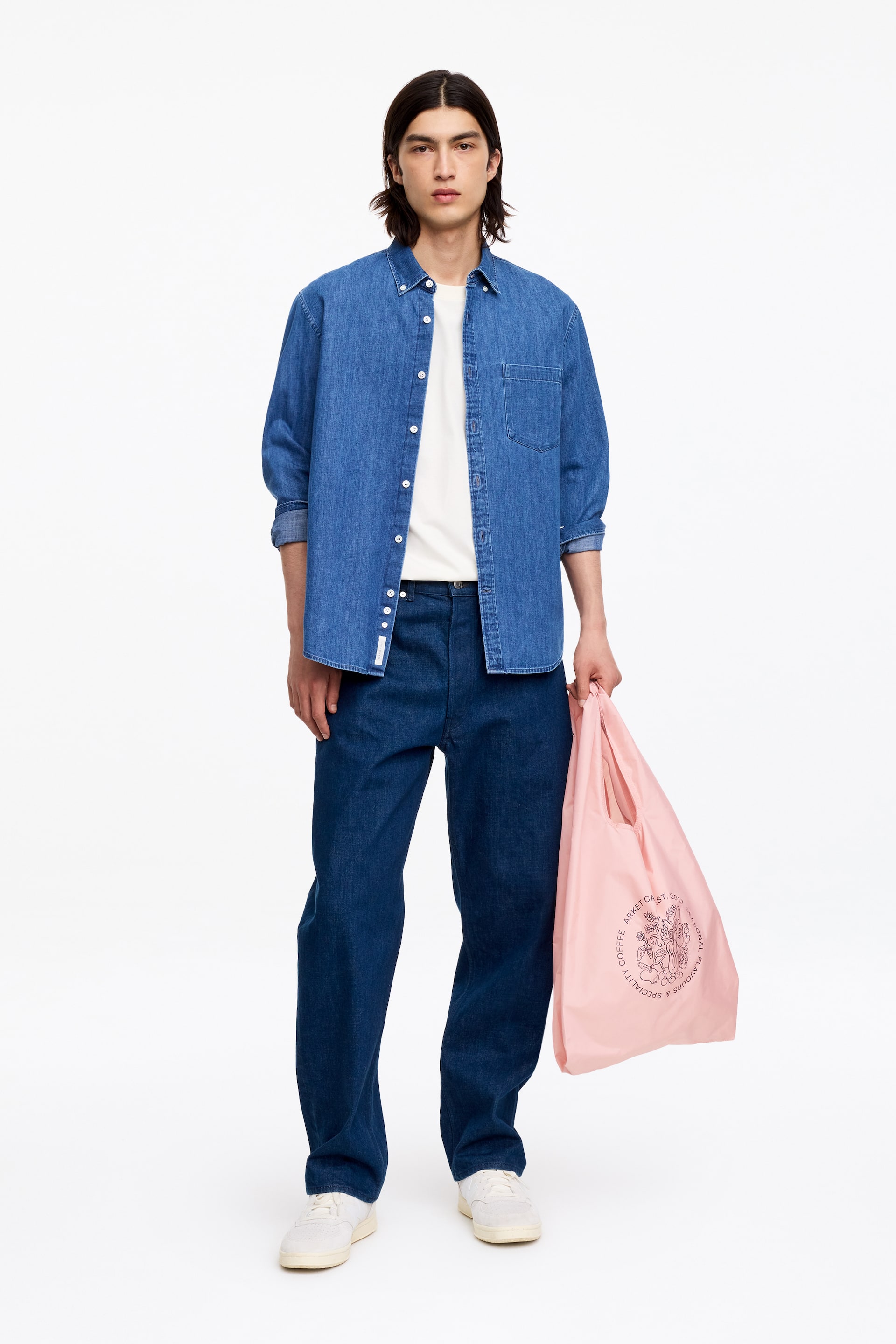 View larger image: The blue denim shirt features a button-down collar and white buttons down the front, with a single patch pocket on the left chest. It is worn open over a white t-shirt, with its long sleeves rolled up, and is facing forward.