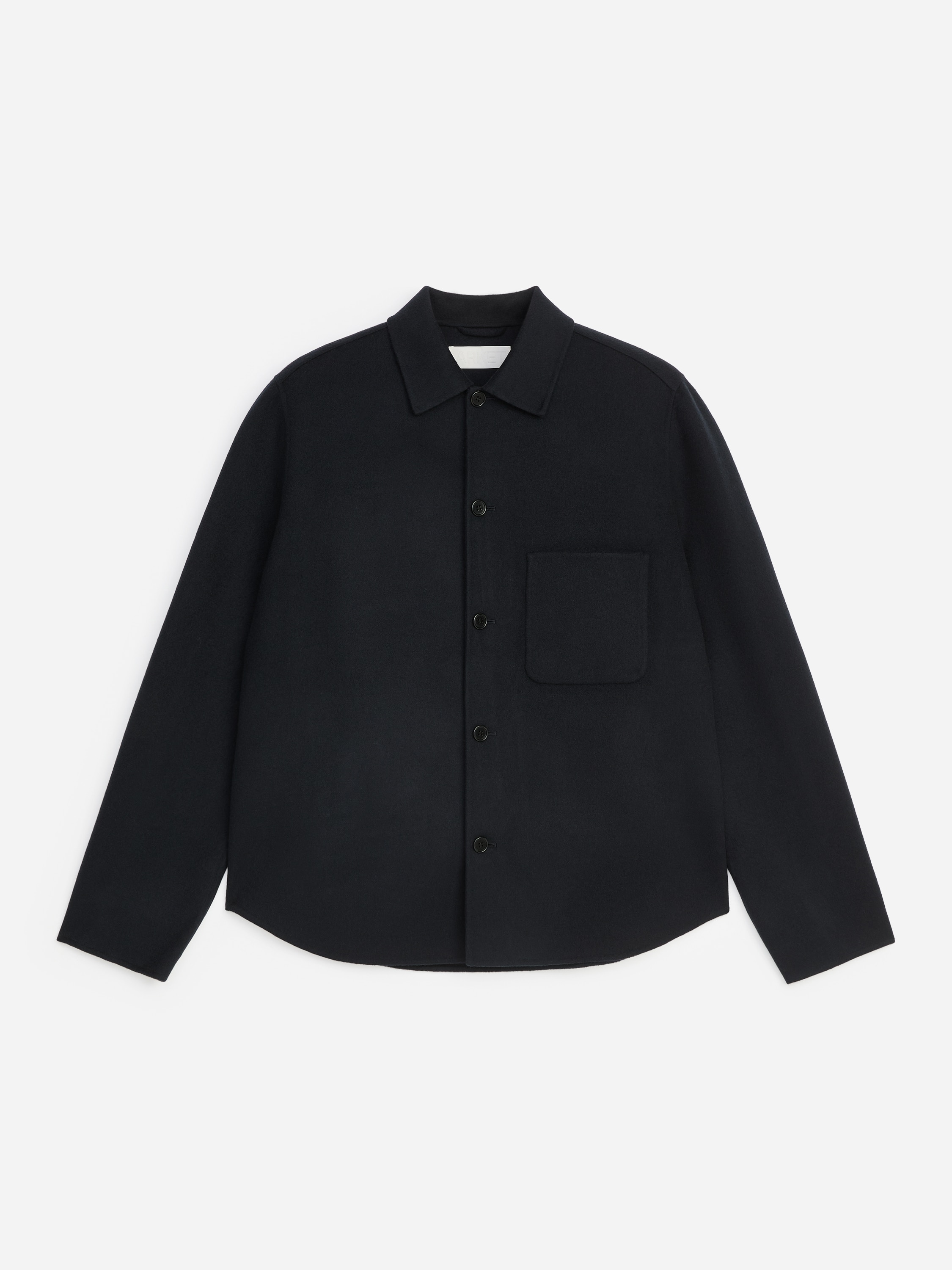 Uma sobrecamisa preta em mistura de lã, com gola clássica e um único bolso de chapa no peito, é usada aberta pela modelo. A sobrecamisa está coordenada sobre uma camisola de malha cinzenta e jeans azuis para evidenciar o seu corte descontraído e regular.