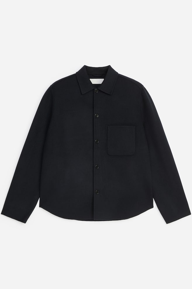 Denne mørk navy overshirt vender front og fremviser et struktureret design med spids krave, frontlukning med fem knapper og en enkelt påsyet brystlomme på venstre side. Plagget har lange ærmer og en diskret buet kant. - 1