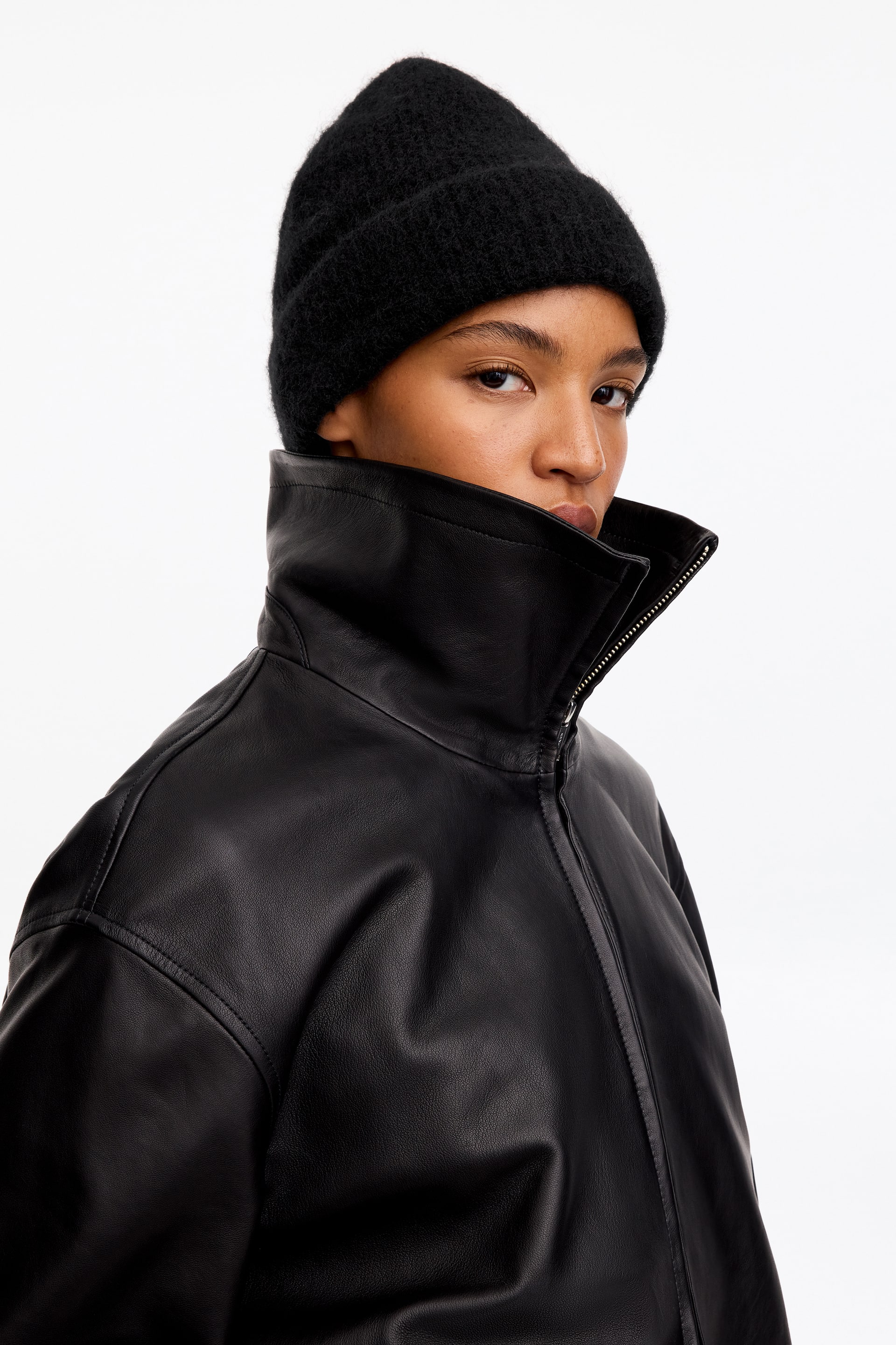 Grotere afbeelding bekijken: Een zwarte gebreide beanie met een gestructureerde, pluizige afwerking en een omgeslagen boord wordt gedragen door het model. De beanie wordt gestyled met een zwart leren jack met hoge kraag, dichtgeritst tot aan de kin.