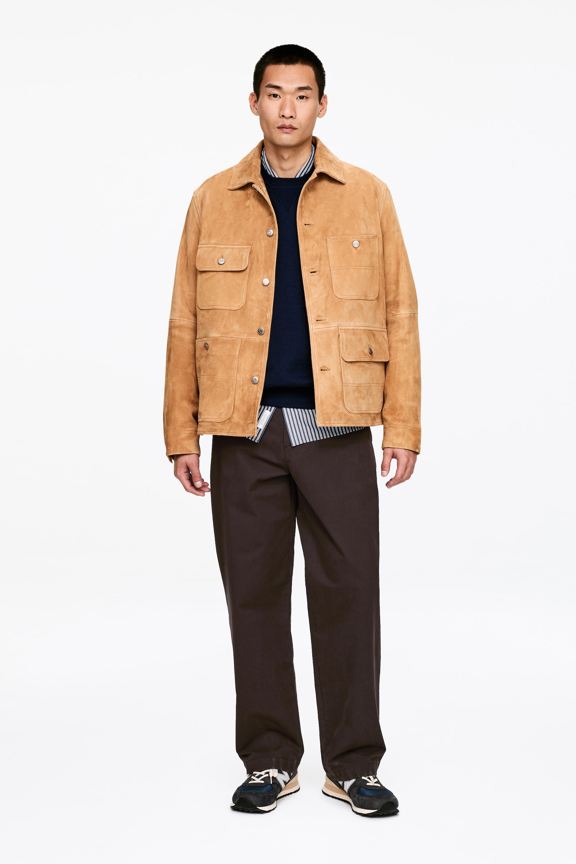 Vezi imaginea mai mare: O jachetă din piele întoarsă maro deschis este prezentată cu fața spre cameră, având un guler, o închidere cu nasturi în față, două buzunare cu clapă la piept și două buzunare aplicate în partea inferioară. Jacheta este stilizată peste un pulover navy cu guler rotund și o cămașă cu dungi, completată de pantaloni maro închis și adidași închiși la culoare.