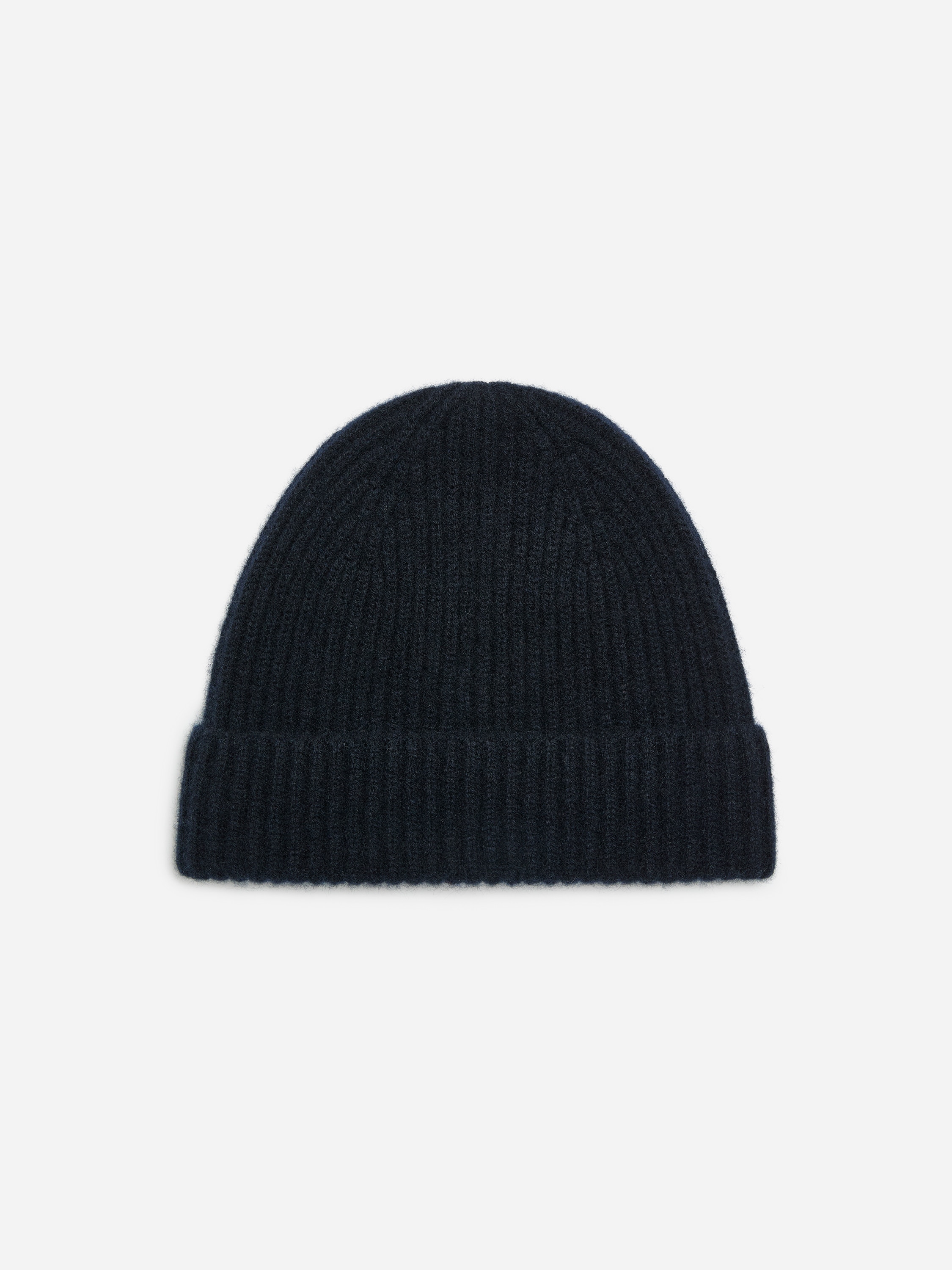En mÃ¶rk marinblÃ¥ stickad beanie med ribbad textur och uppvikt kant bÃ¤rs av modellen. BeaniemÃ¶ssan stylas med en mÃ¶rk marinblÃ¥ kappa som bÃ¤rs ovanpÃ¥ en klarblÃ¥ rundhalsad trÃ¶ja.