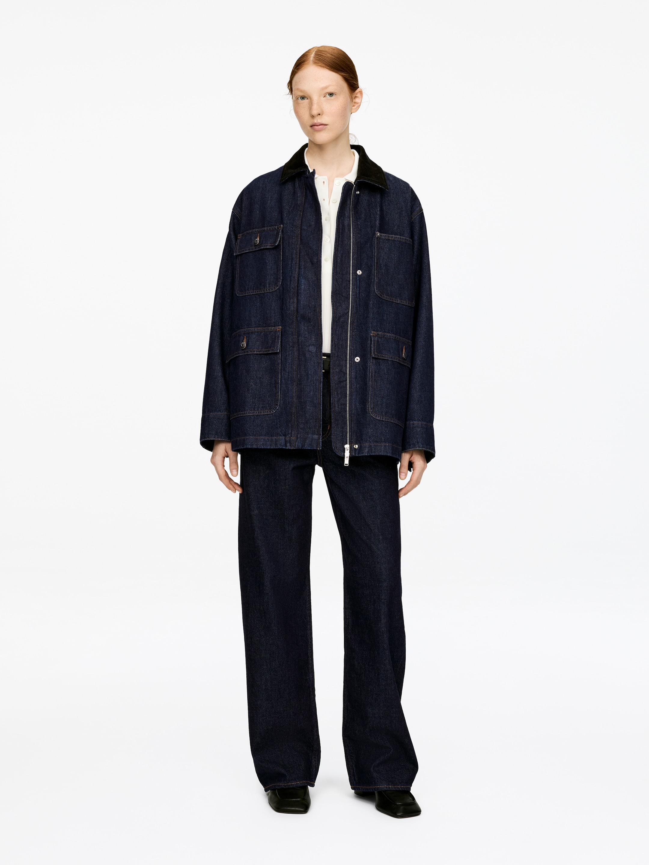 Visualizza immagine più grande: Una giacca di jeans oversize blu scuro con un colletto di velluto a coste nero è indossata dal modello. La chiusura con zip centrale anteriore con pattina è parzialmente aperta, rivelando quattro tasche applicate frontali con pattine e bottoni, mentre il modello la indossa sopra una camicia bianca e pantaloni scuri.