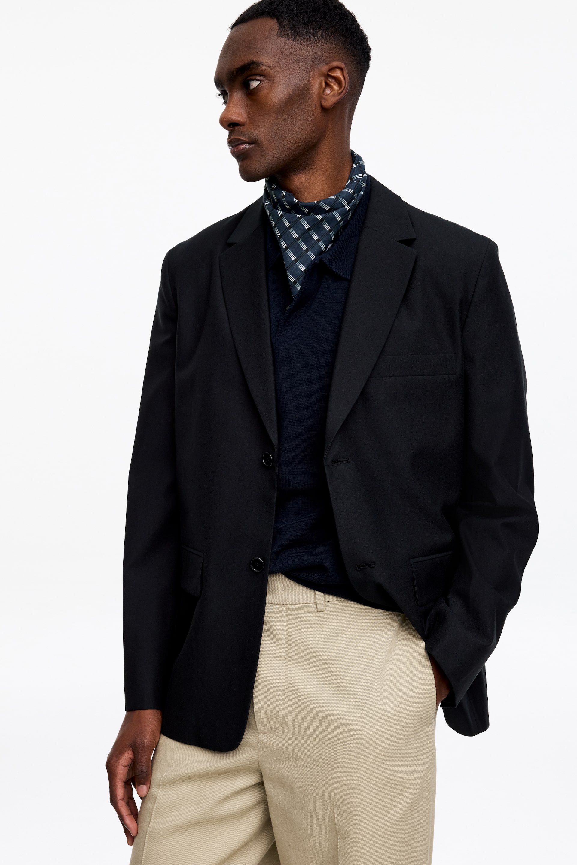 Agrandir l'image: Un blazer noir est présenté sous un angle légèrement frontal-droit, avec des revers crantés, une fermeture à deux boutons, une poche poitrine passepoilée et deux poches à rabat. Il est superposé sur un polo bleu marine foncé, avec un foulard à motifs visible au niveau du cou, et assorti à un pantalon beige clair.