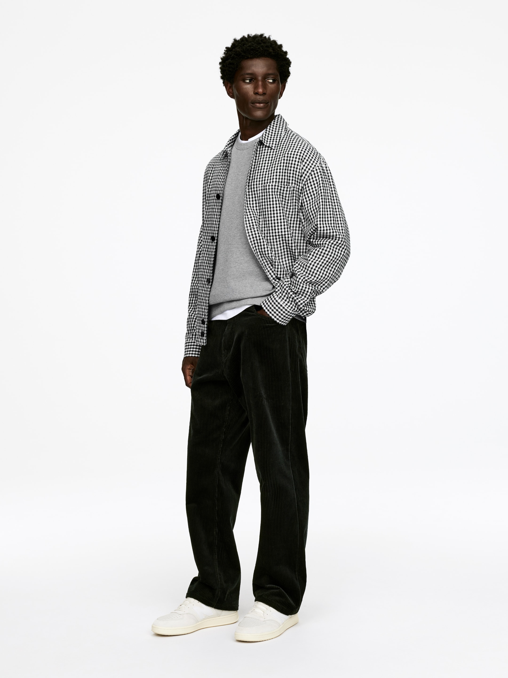 Ingrandisci l'immagine: Una camicia a quadri in gingham bianco e nero viene indossata aperta, stratificata su una felpa grigia con scollo rotondo e una t-shirt bianca. Pantaloni a coste larghe neri, con una vestibilità rilassata e dritta, sono abbinati a sneakers basse color bianco sporco.
