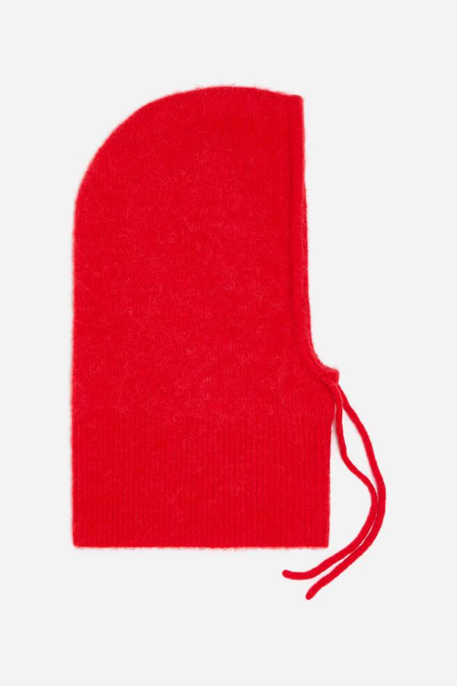 A bright red knitted balaclava is shown in side profile, facing right, featuring a fuzzy texture and a ribbed lower neck. Two thin red knitted drawstrings hang from the front chin opening. - 1
