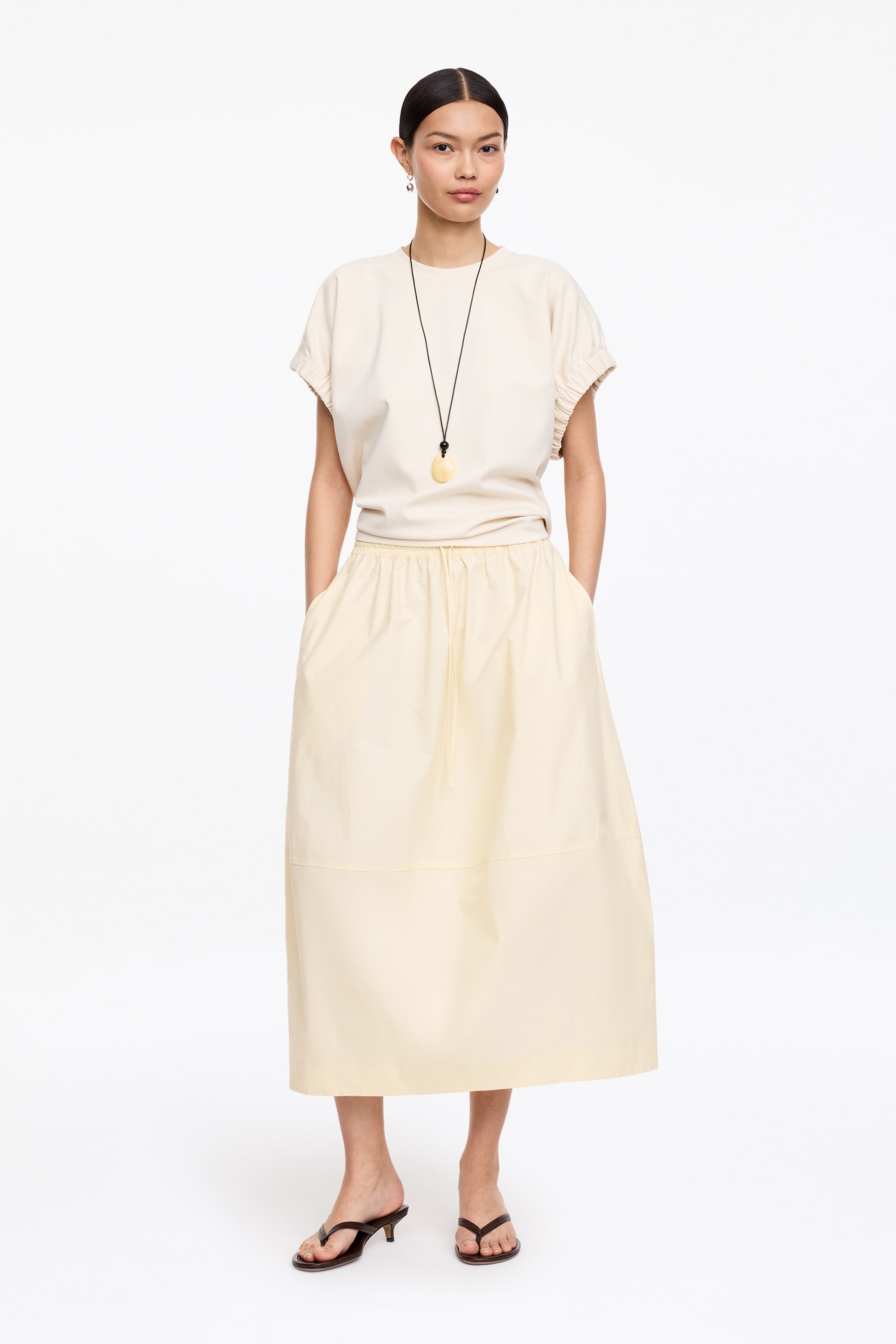 Ver imagen más grande: Una blusa de cuello redondo en color crema pálido con mangas cortas fruncidas se lleva metida en una falda midi amarilla pálida que presenta una cintura con cordón ajustable y un corte holgado. Un collar largo de cordón negro con un colgante ovalado ligero se superpone sobre la blusa.