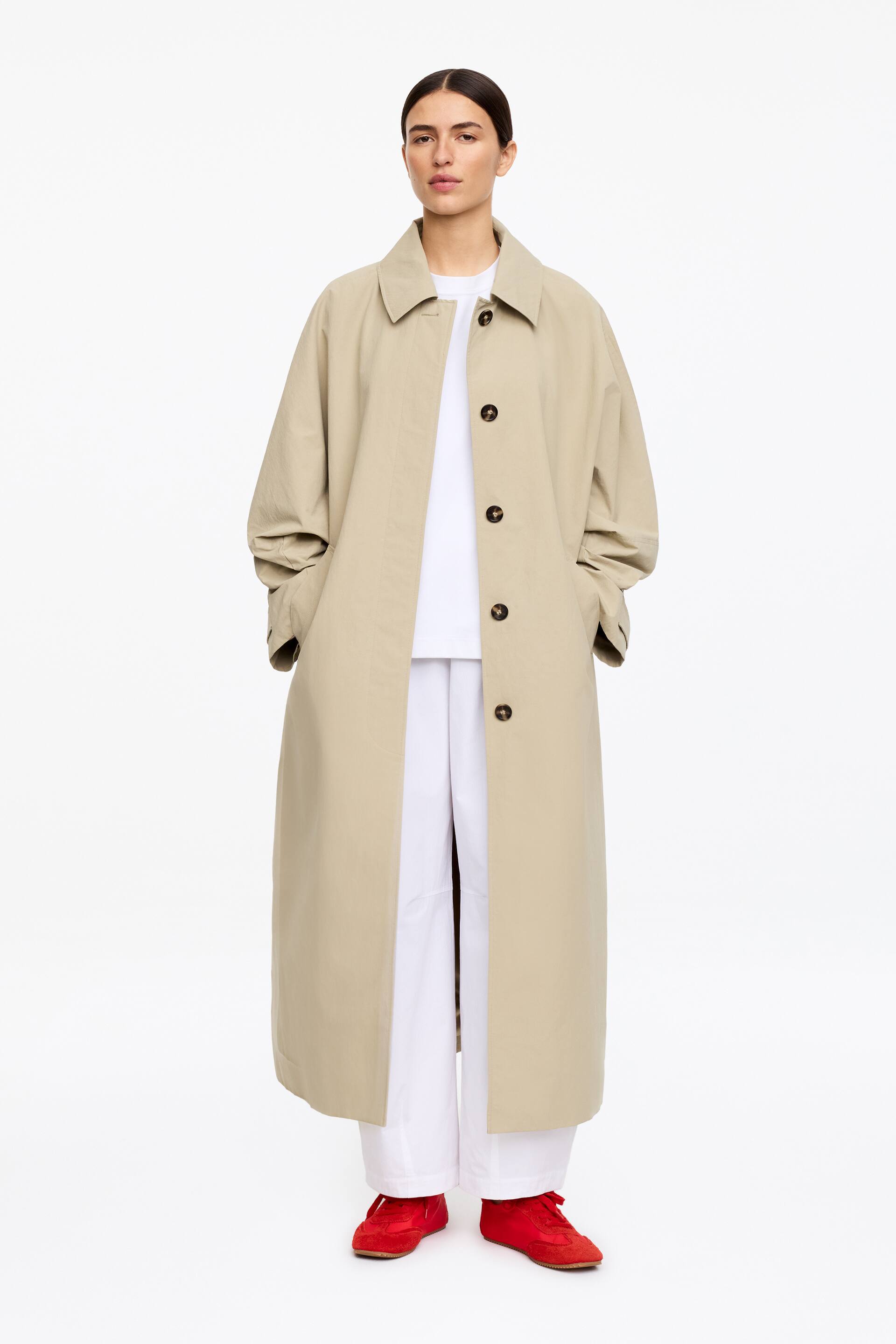 Ver imagem maior: Um casaco comprido bege apresenta um corte descontraído e de comprimento total sobre uma t-shirt branca e calças brancas. Apresenta uma gola aberta, um fecho de botões simples com botões castanho-escuros, mangas compridas com punhos franzidos e bolsos laterais.