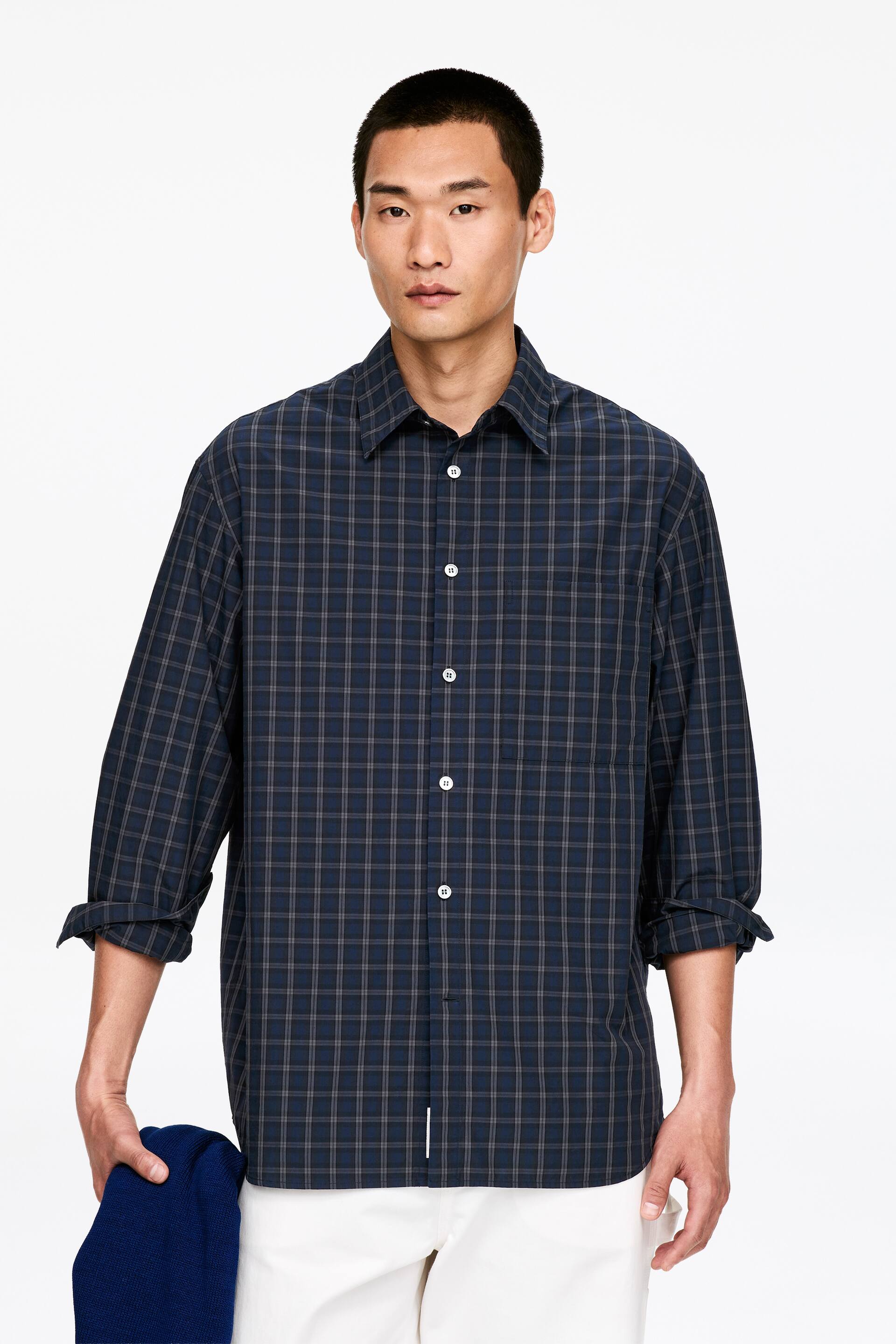 Agrandir l'image: Une chemise à carreaux bleu foncé est présentée de face, affichant un motif en grille de fines lignes beige et blanches. Elle est dotée d'un col classique à pointes, d'une fermeture à boutons avec des boutons blancs, et d'une poche poitrine plaquée unique. Ses manches longues sont retroussées.